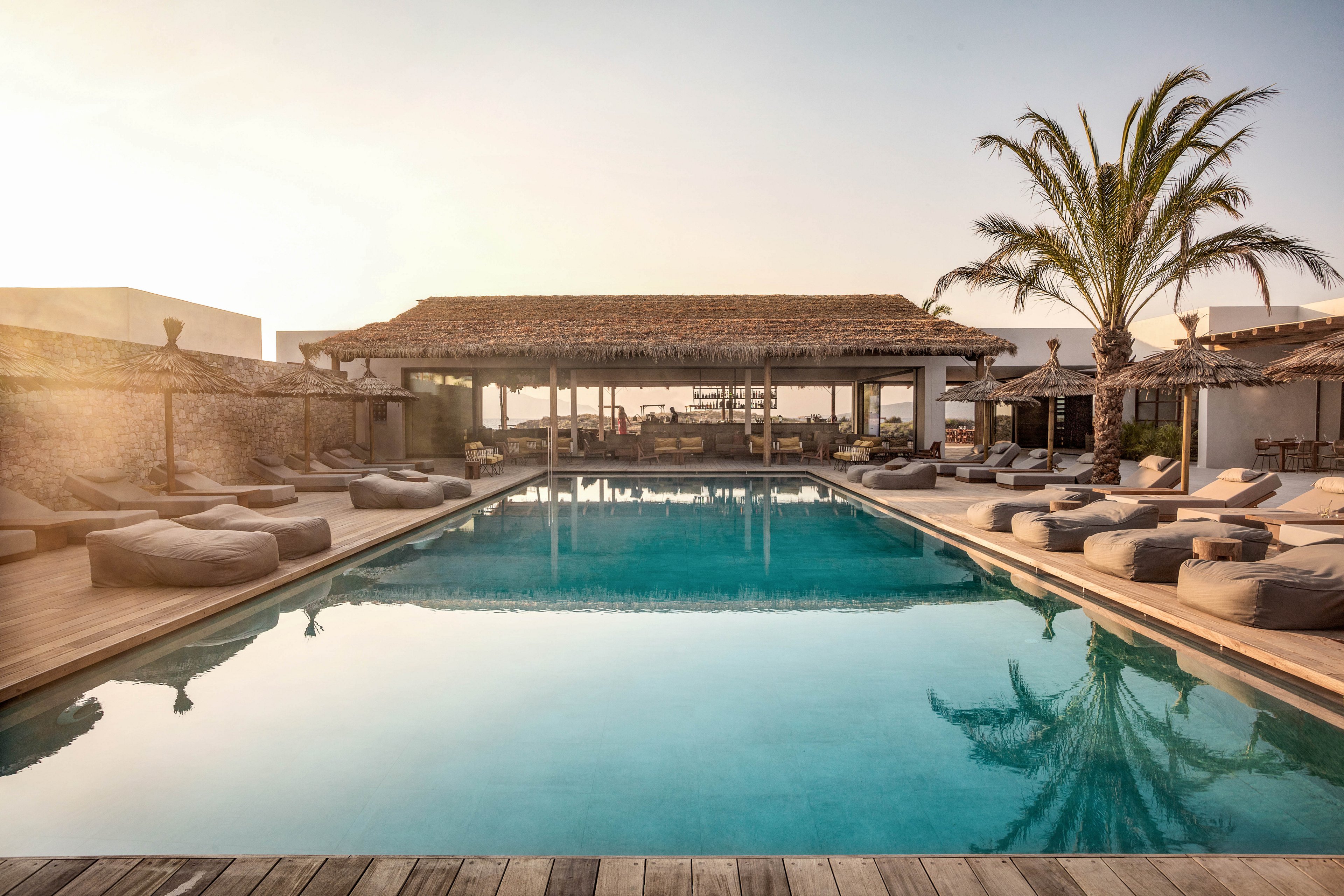 The image features a serene outdoor swimming pool surrounded by cushioned lounge chairs and bean bags. The setting is a modern resort or villa, with a rustic wooden pavilion and palm trees adding to the tropical ambiance. The lighting is warm and golden, suggesting late afternoon or sunset, and the water reflects the tranquil mood. No visible text or numeric values are present in the image.