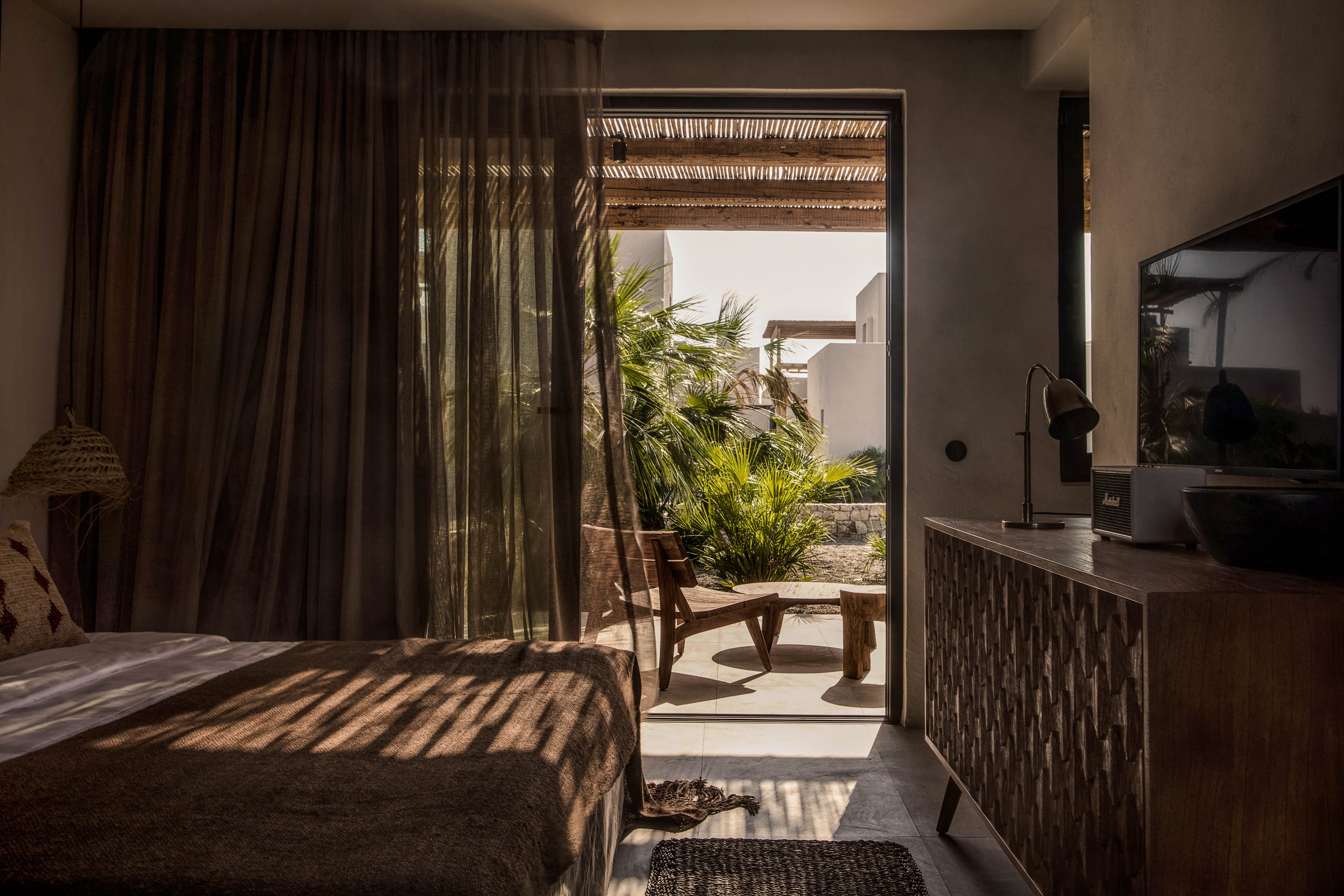 A contemporary bedroom features a cozy bed, textured curtains, and a wooden dresser. Sunlight streams through the sheer curtain, casting shadows across the room and highlighting the outdoor patio with lush tropical plants and wooden furniture. The neutral palette and natural materials create a warm, inviting atmosphere, blending indoor comfort with outdoor serenity.