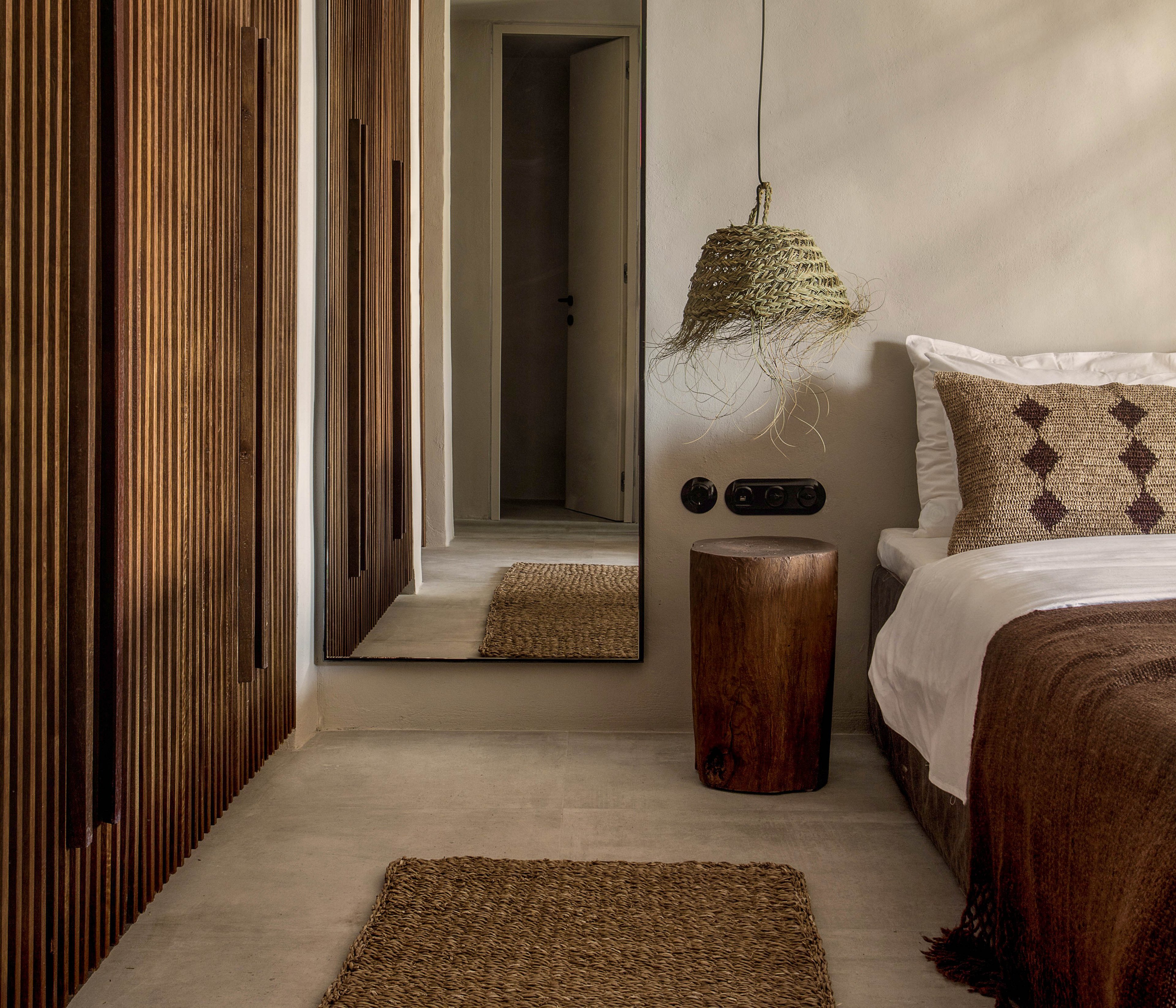 A cozy bedroom features a minimalist design with natural wood paneling and woven elements. The space includes a bed with a geometric-patterned pillow, a woven rug, and a rustic wooden stool. A hanging lamp with a natural fiber shade adds texture, while a large mirror reflects the neutral palette. The overall mood is warm and earthy, with brown and beige tones dominating the scene.