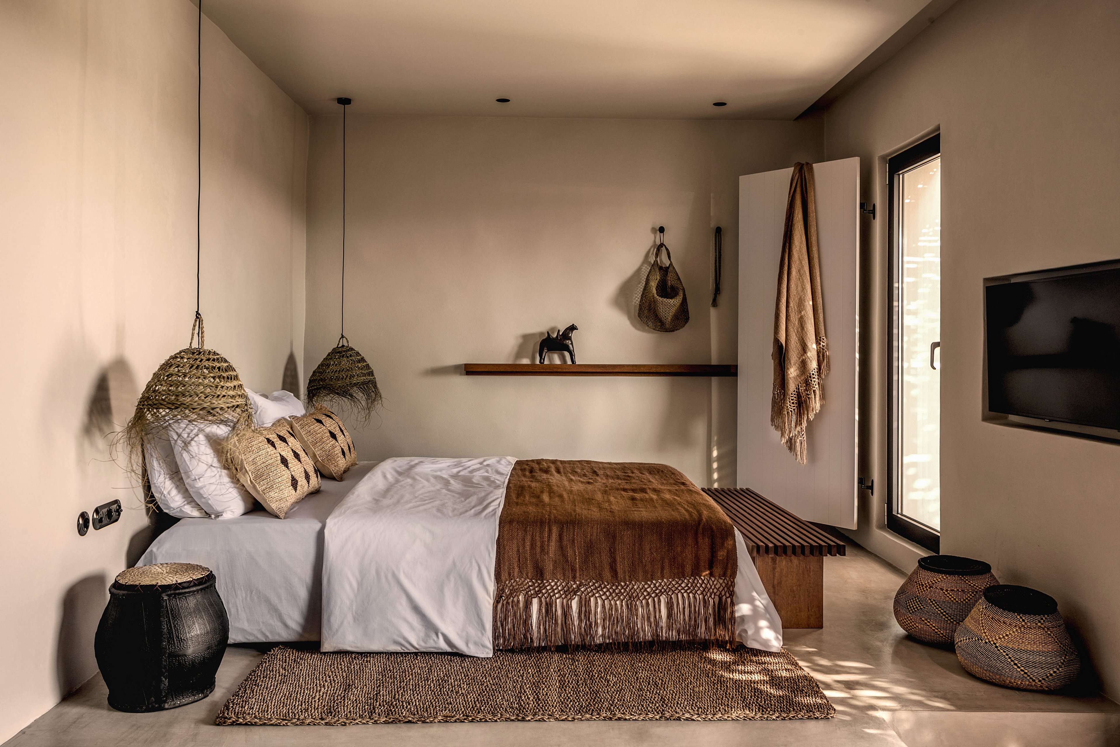 A cozy bedroom features a neatly made bed with white and brown linens, surrounded by woven baskets and hanging lamps. The space is styled with earthy tones and natural textures, including a jute rug and wooden bench. Soft lighting and minimalist decor create a warm, inviting atmosphere. No visible text or numeric values are present in the image.