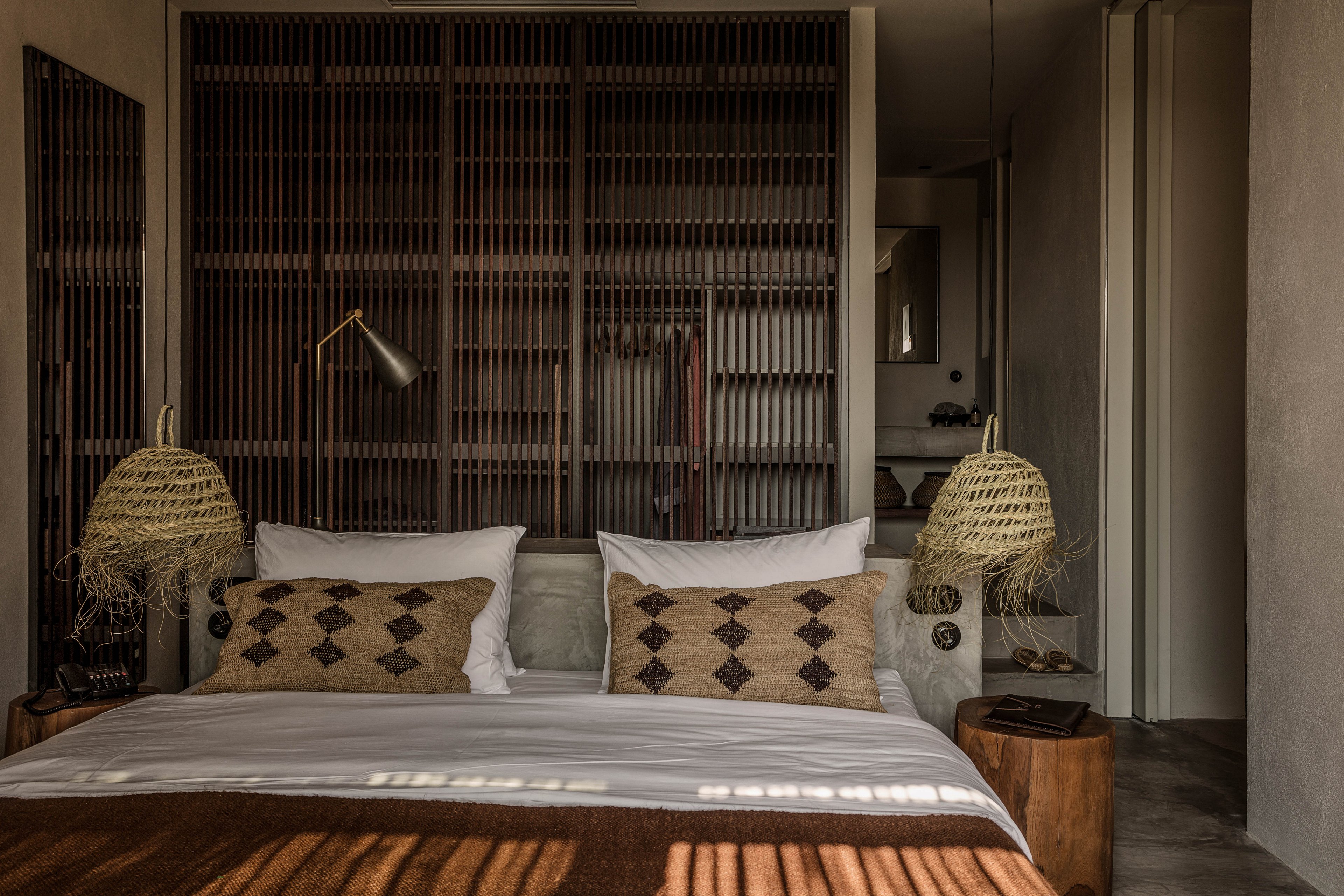 A cozy bedroom features a large bed with geometric-patterned pillows and a brown throw blanket. The room is styled with woven pendant lamps on either side, wooden side tables, and a dark slatted partition behind the bed. Warm, earthy tones and natural textures create a calm, inviting atmosphere.