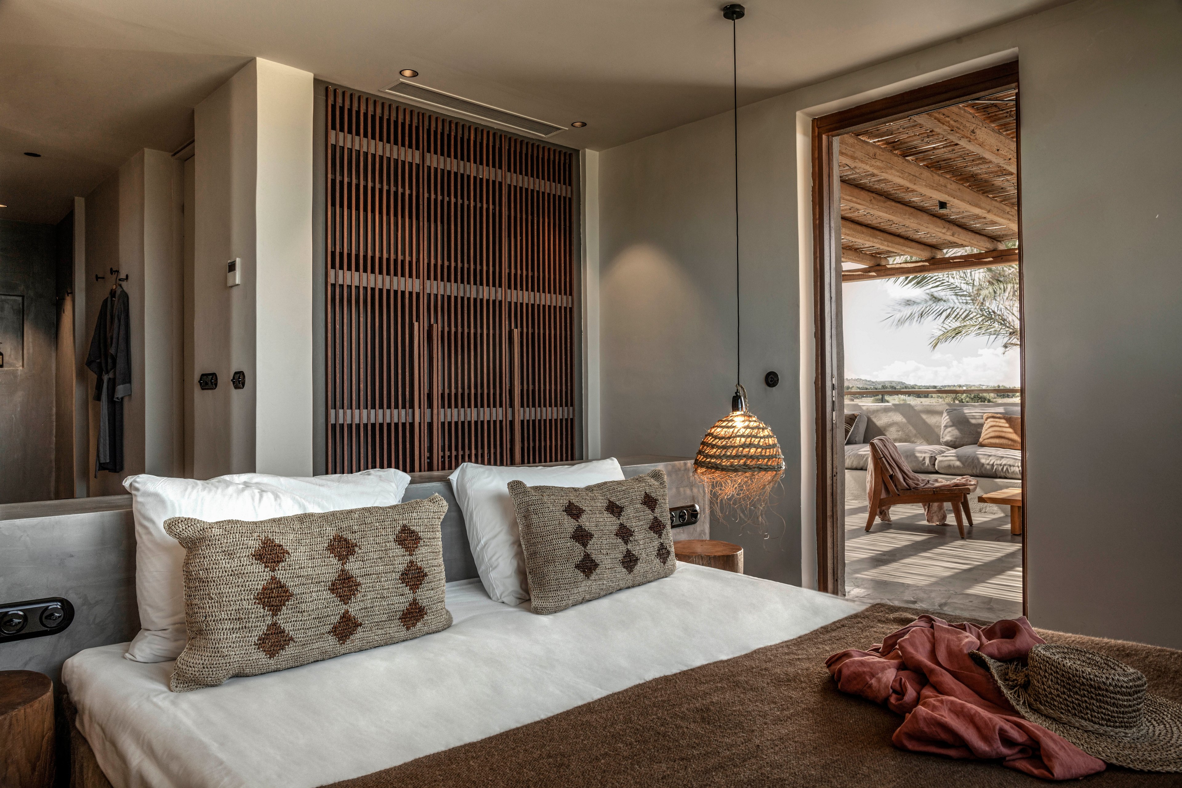 A contemporary hotel room features a neatly made bed with textured pillows and a brown blanket. The space is illuminated by a woven pendant lamp, and a wooden accent wall adds warmth. Through the open door, an inviting outdoor patio with lounge chairs and natural decor is visible, creating a relaxed atmosphere. Earthy tones and soft lighting contribute to a cozy, tranquil mood.