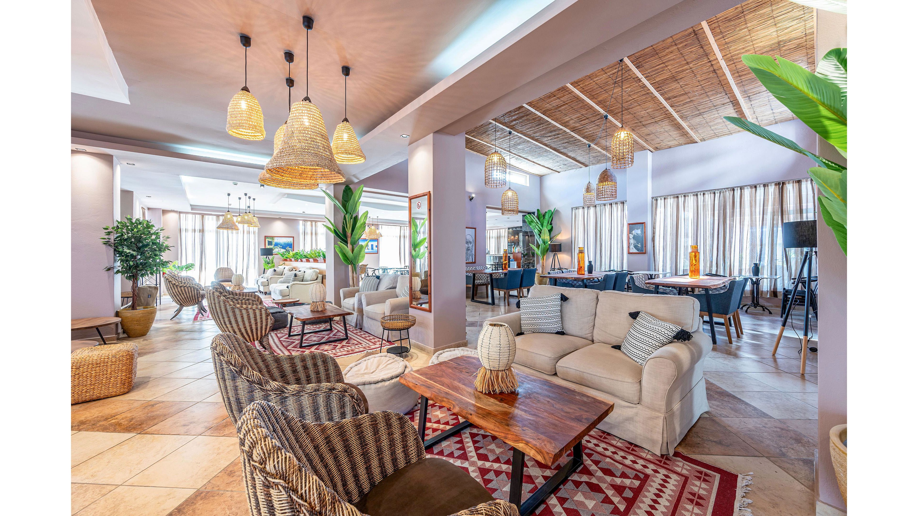 Spacious lounge area featuring comfortable sofas and wicker armchairs arranged around wooden coffee tables. The setting appears to be a modern hotel or resort lobby with large windows and sheer curtains letting in natural light. Warm pendant lights hang from a wooden ceiling, and patterned rugs add color and texture to the tiled floor. Green potted plants and decorative baskets enhance the relaxed, coastal-inspired atmosphere.