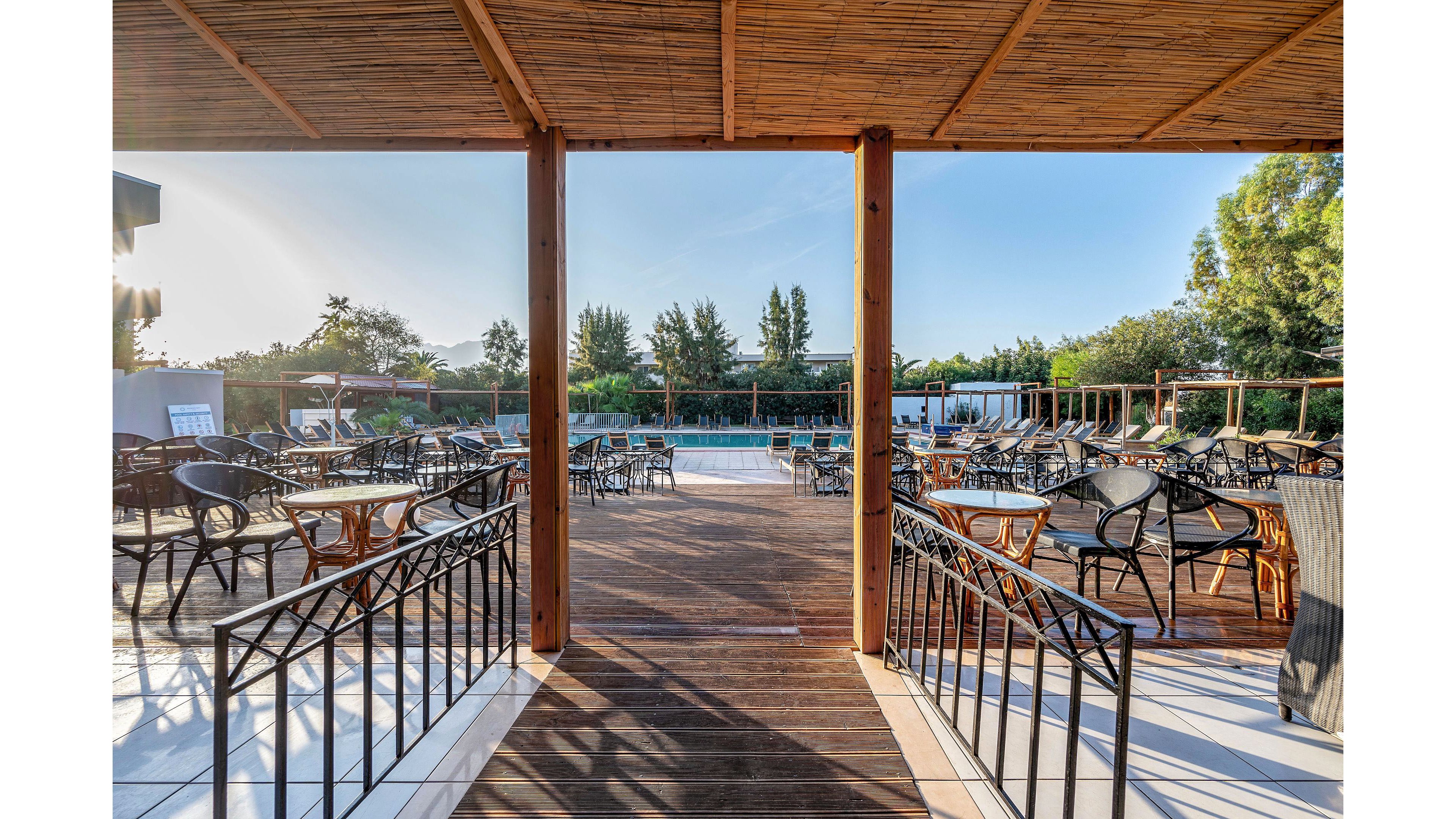 An open-air café terrace overlooks a swimming pool surrounded by greenery under a clear sky. Empty tables and wicker chairs are neatly arranged on a wooden deck framed by a pergola structure. Metal railings guide the view toward the pool area, creating a symmetrical composition. The warm morning light and natural materials give the scene a relaxed, inviting atmosphere.