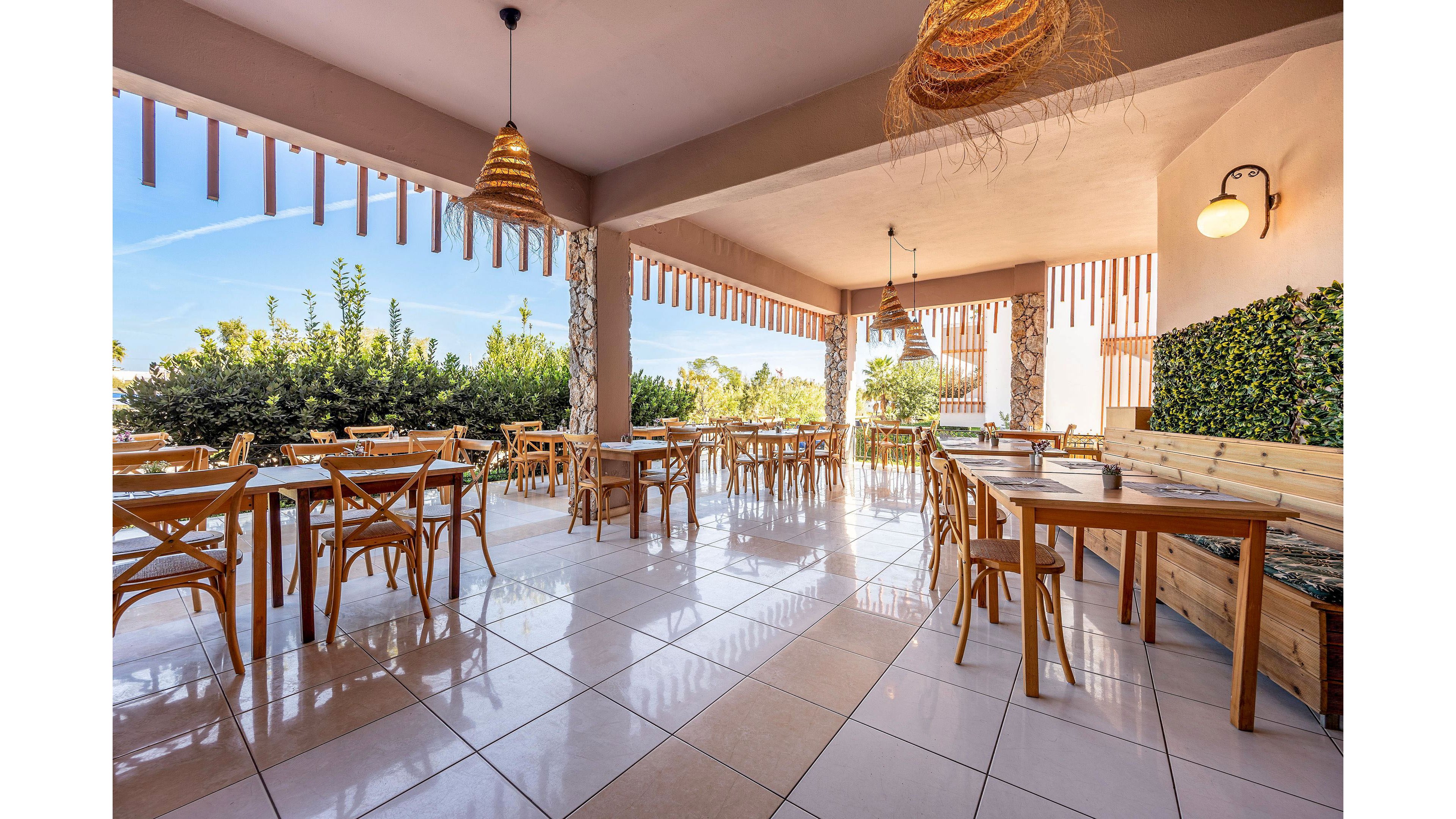 Spacious outdoor dining area with multiple wooden tables and chairs arranged neatly on a tiled floor. The terrace is covered by a roof with hanging woven light fixtures and decorative vertical slats along the edges. Green shrubs and plants surround the space, creating a natural, relaxed atmosphere. Warm sunlight enhances the inviting, casual feel of the restaurant setting.