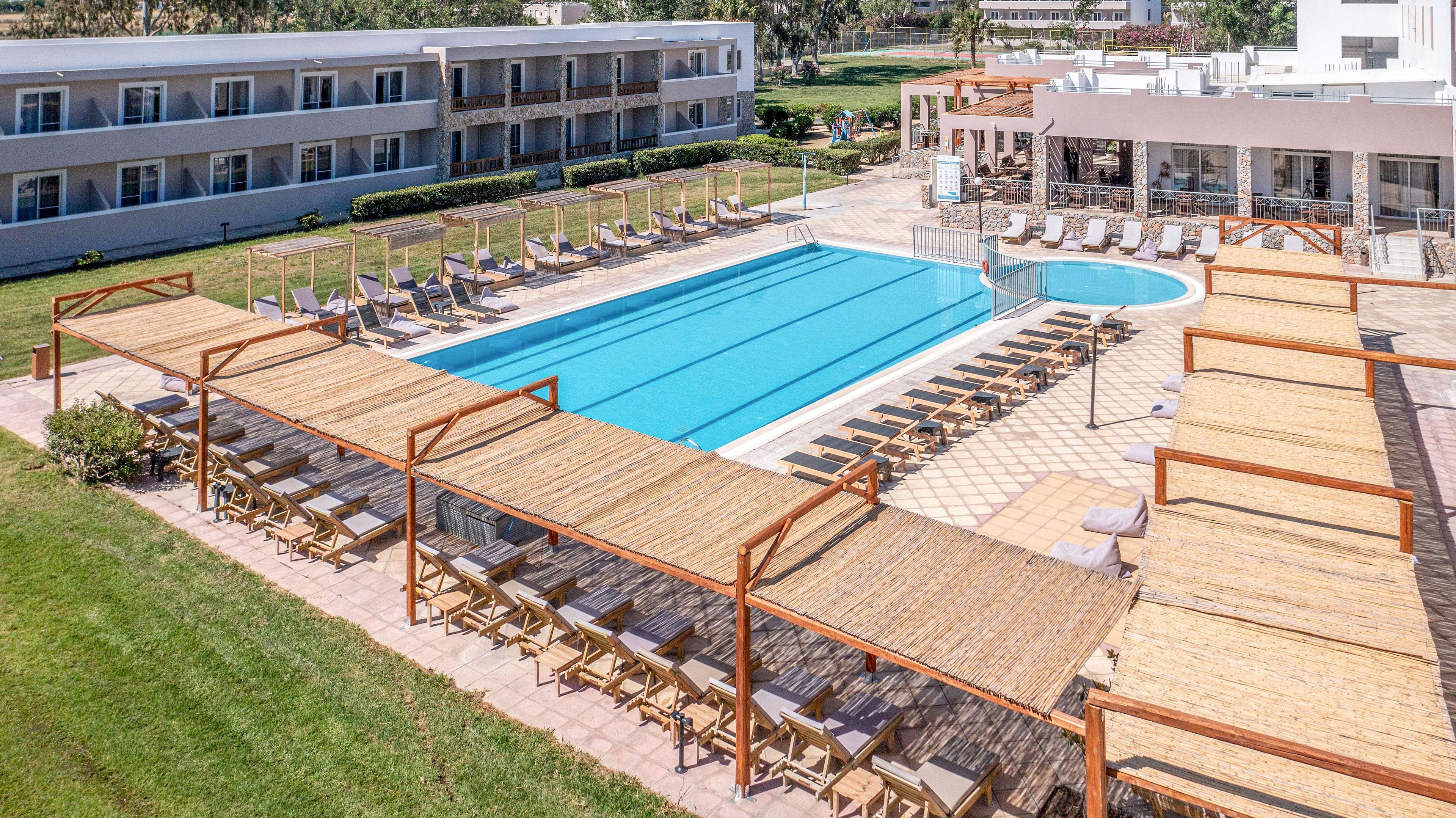 A large rectangular outdoor swimming pool is surrounded by rows of sun loungers and umbrellas at a modern resort. Wooden pergolas with shaded seating and tables line the foreground, creating a comfortable relaxation and dining area. A smaller circular pool sits adjacent to the main pool near a white building with large windows. In the background, low-rise hotel buildings with balconies overlook the pool area, all set in a bright, sunny atmosphere with green lawns.