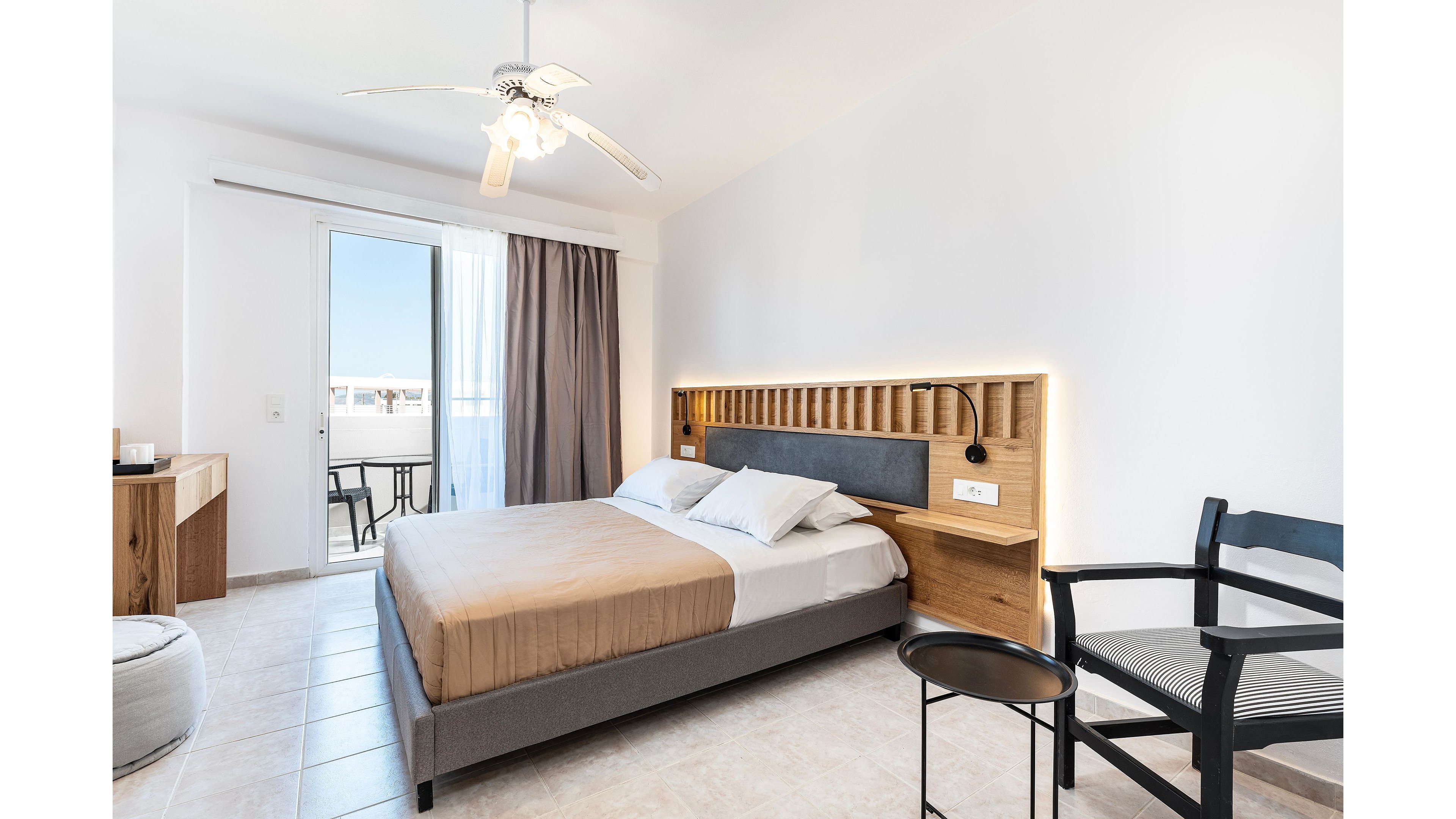 Contemporary hotel room featuring a double bed with neutral bedding and a wood-accent headboard. The space is bright and airy, with white walls, tiled floor, and a ceiling fan providing comfort. A glass door opens to a small balcony with outdoor chairs and a table, letting in natural light. Minimalist furniture, including a desk, chair, and side table, creates a clean and relaxing atmosphere.