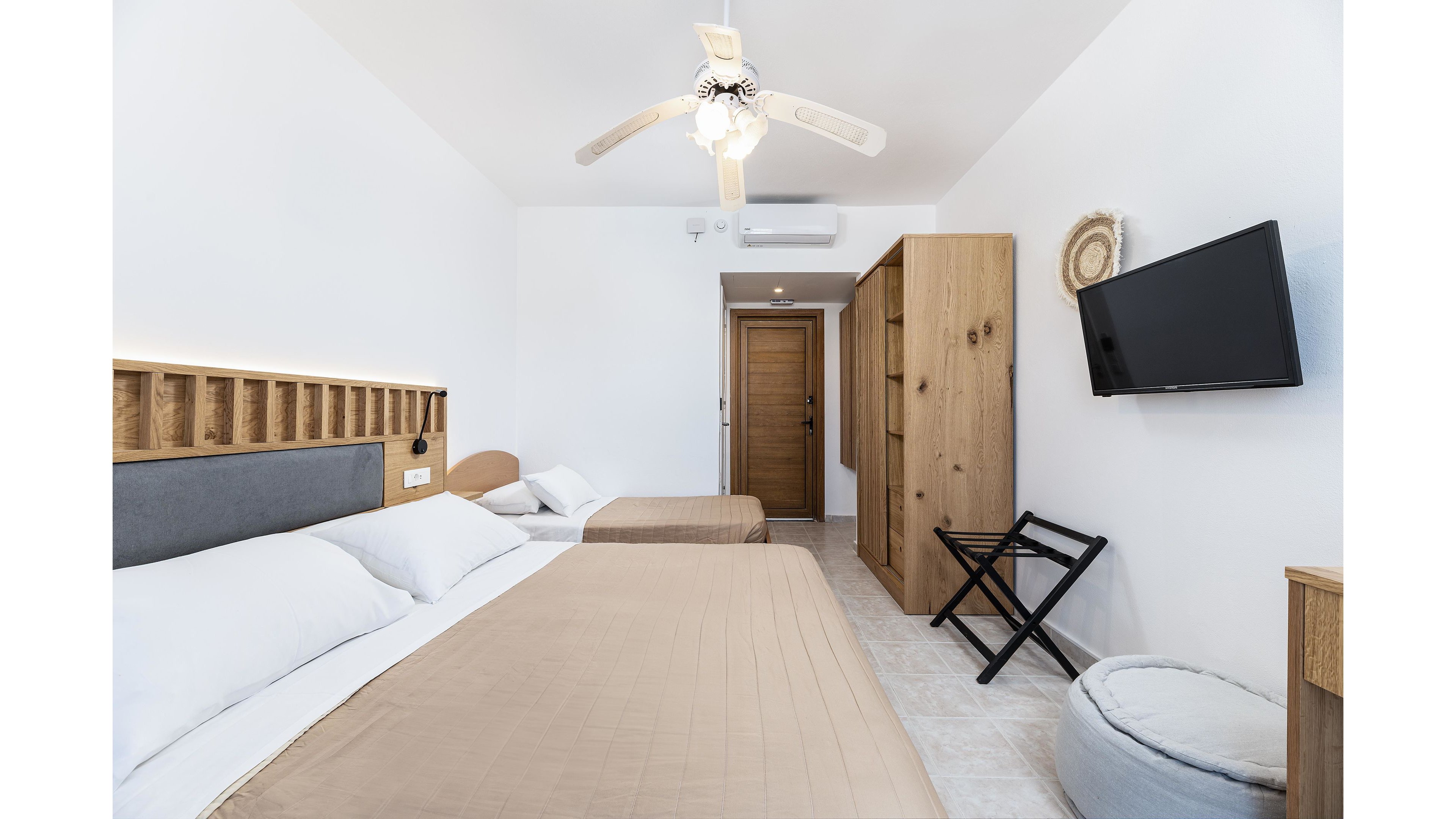 A clean, modern hotel room features a large double bed and a smaller single bed with neutral beige bedding. The space is bright with white walls, a ceiling fan, and tiled flooring. A wooden wardrobe and door add warmth, while a wall-mounted flat-screen TV faces the beds. Additional furnishings include a folding luggage rack and a round upholstered ottoman, creating a simple yet comfortable guest room.