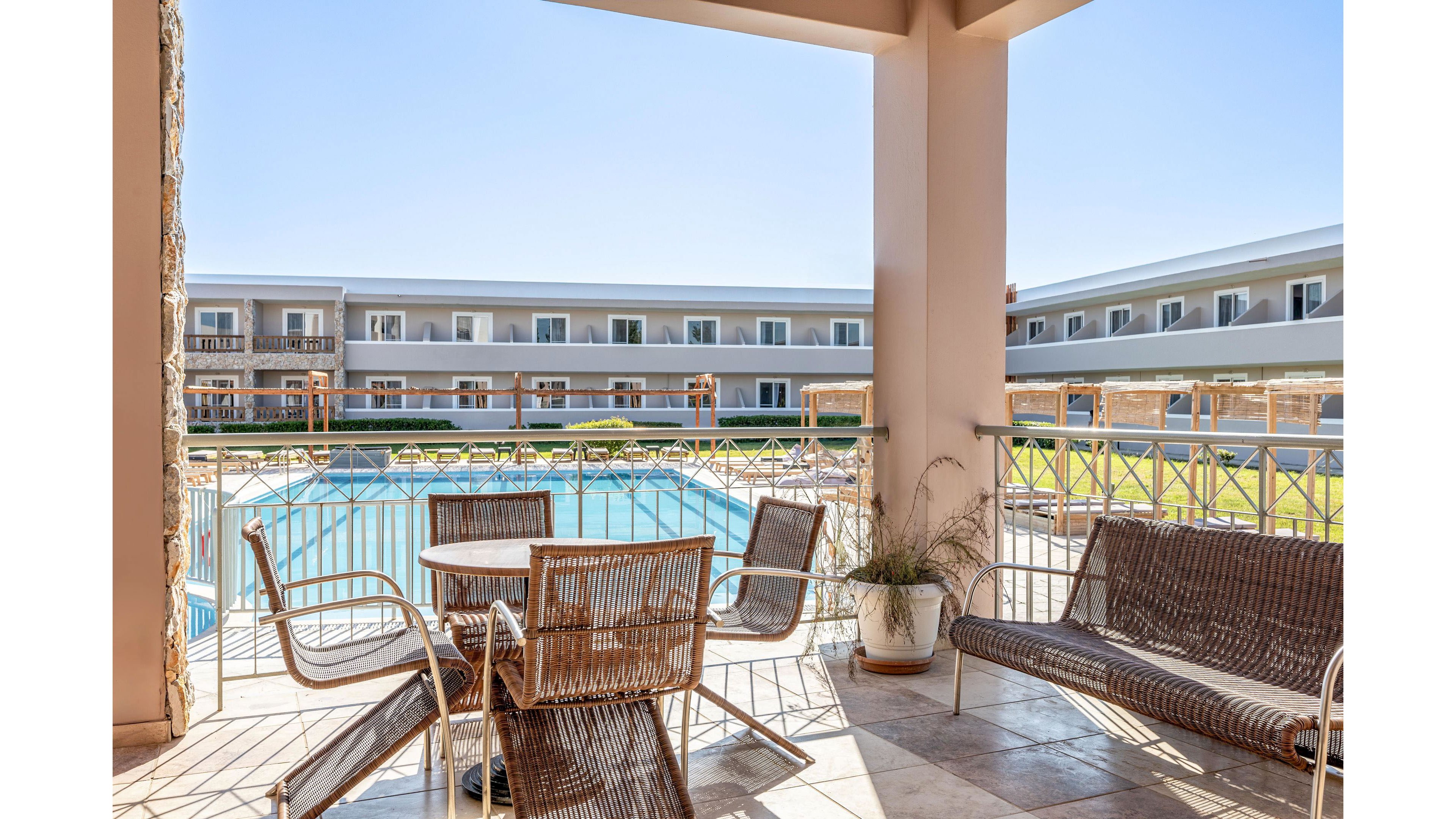 An outdoor terrace with wicker chairs and benches overlooks a large rectangular swimming pool. The scene is part of a modern resort or hotel complex with two-story buildings surrounding the pool area. Bright sunlight and clear blue sky create a warm, inviting atmosphere. A potted plant and tiled floor add decorative detail to the relaxing seating space.