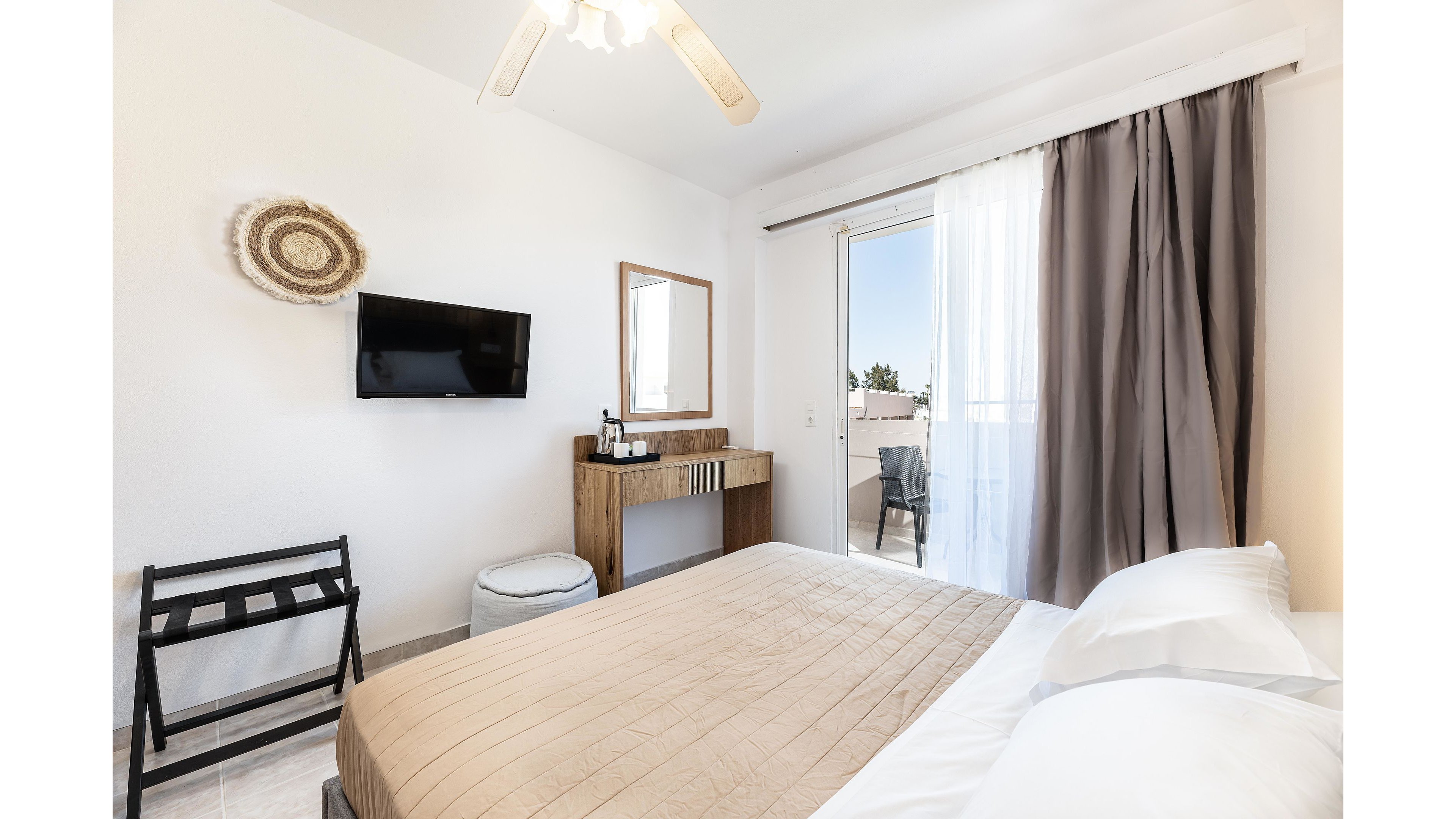 A modern hotel bedroom features a neatly made double bed with beige bedding and white pillows. A wall-mounted flat-screen TV faces the bed, next to a wooden vanity table with a mirror and small amenities. Sheer white curtains and dark drapes frame a glass door that opens to a sunny balcony with outdoor seating. The room is decorated in a light, neutral palette, creating a clean and relaxing atmosphere.