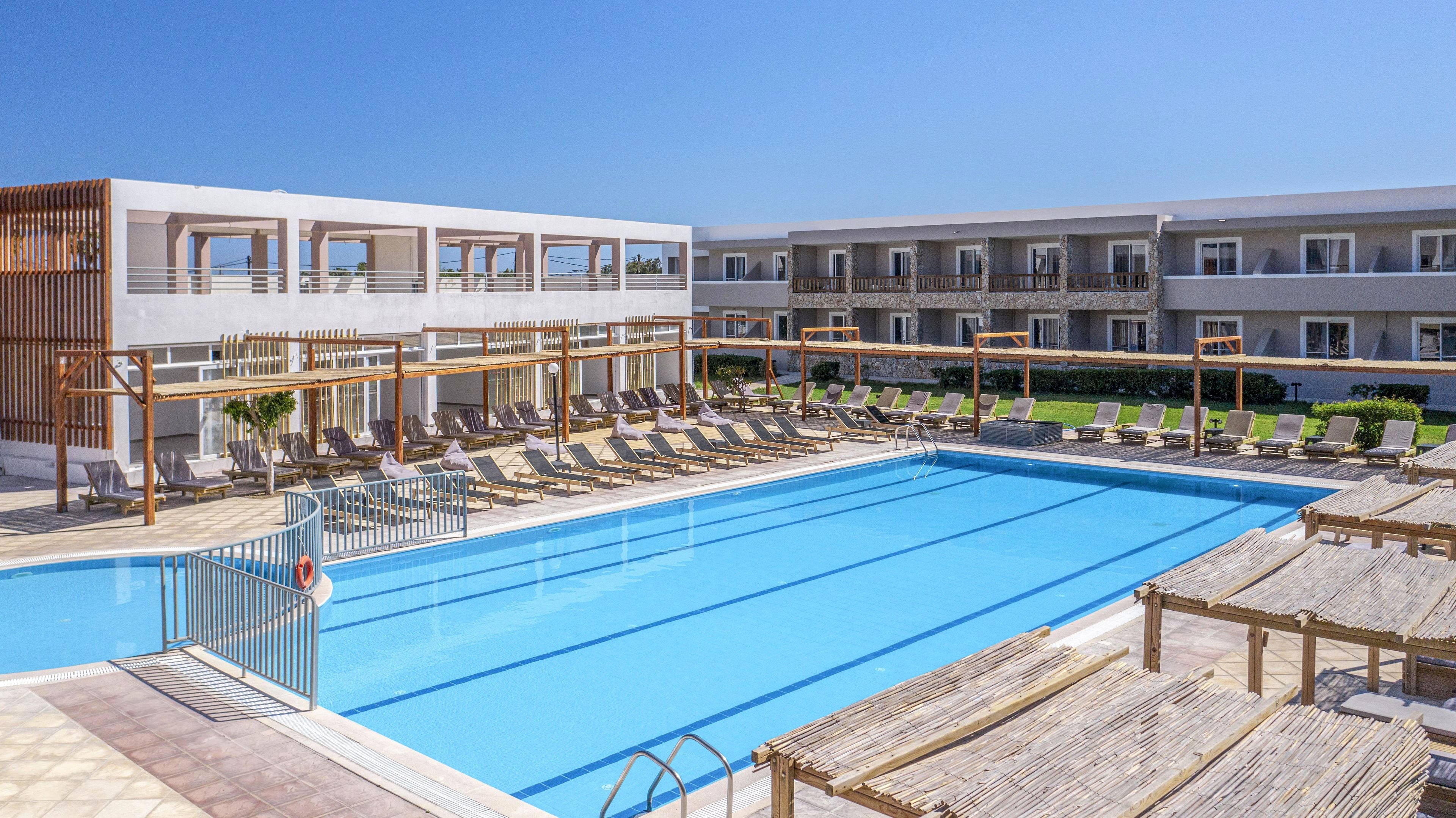 A contemporary resort hotel features a large rectangular outdoor swimming pool surrounded by sun loungers. The setting appears bright and sunny under a clear blue sky, suggesting a warm vacation destination. Two-story white buildings with balconies frame the pool area, while wooden pergolas provide shaded seating. The overall scene conveys relaxation, leisure, and a clean, minimalist architectural style.