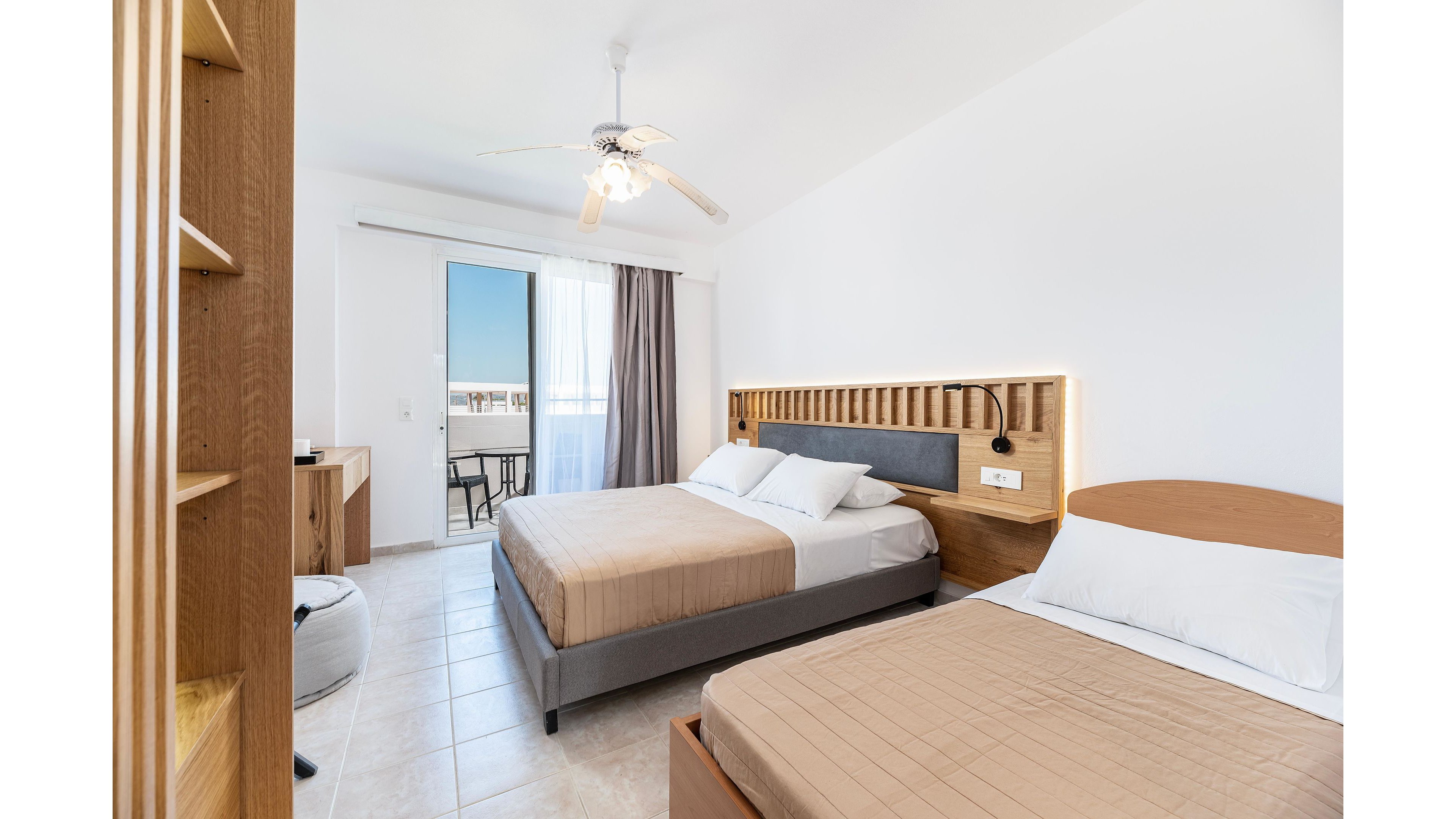A modern hotel room features two neatly made double beds with beige bedding and white pillows. The space is bright and airy, with light-colored tile flooring and white walls. A ceiling fan hangs above, and a desk with chair sits near the sliding glass door. The door opens to a balcony with outdoor chairs and a view of the blue sky and sea.