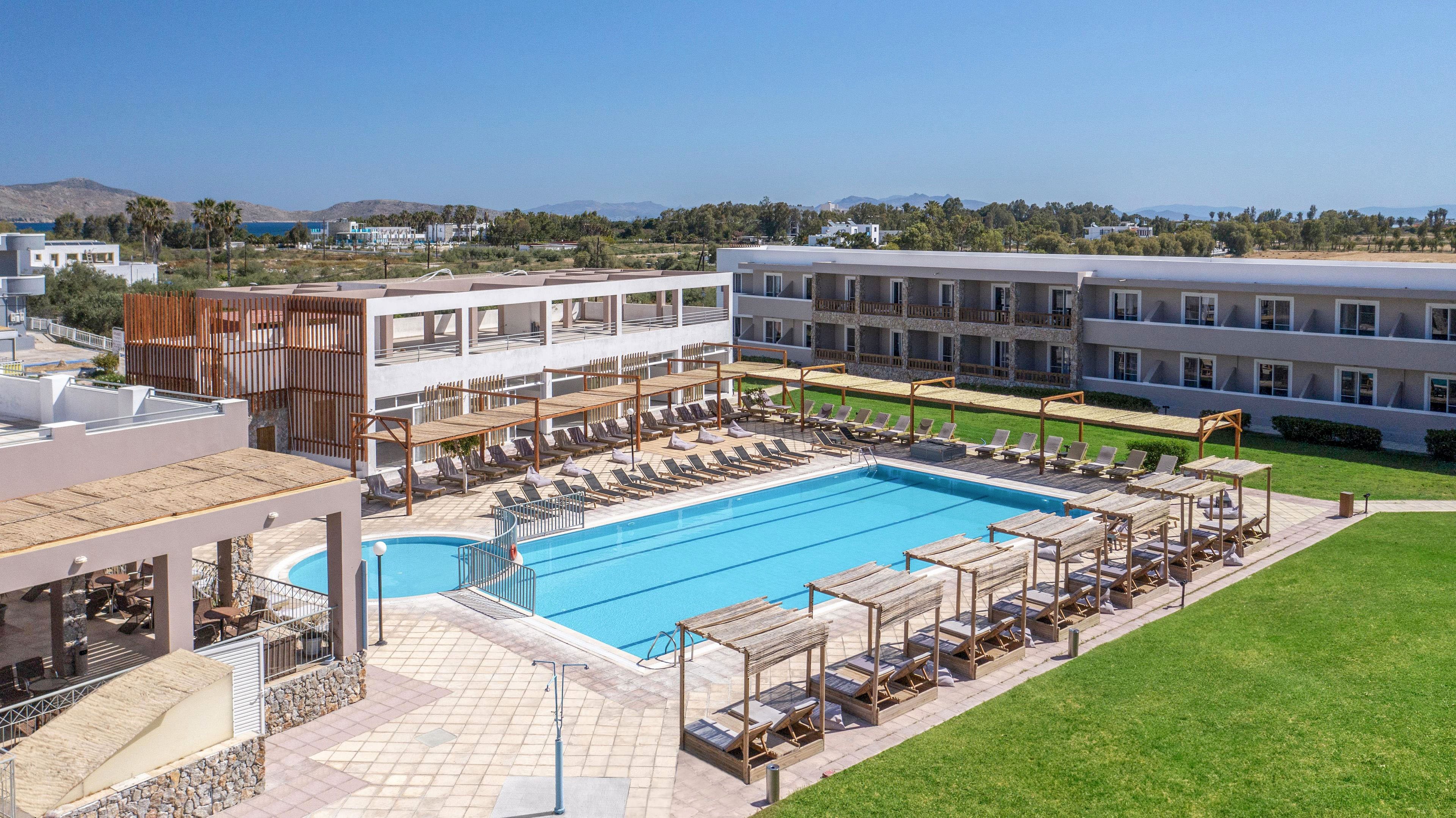 A contemporary resort hotel features a large rectangular outdoor swimming pool surrounded by sun loungers and wooden cabanas. The buildings are low-rise with white facades and balconies overlooking the pool area. Clear blue sky and distant greenery suggest a warm, sunny vacation destination. The scene conveys relaxation, leisure, and holiday atmosphere in a well-maintained property.