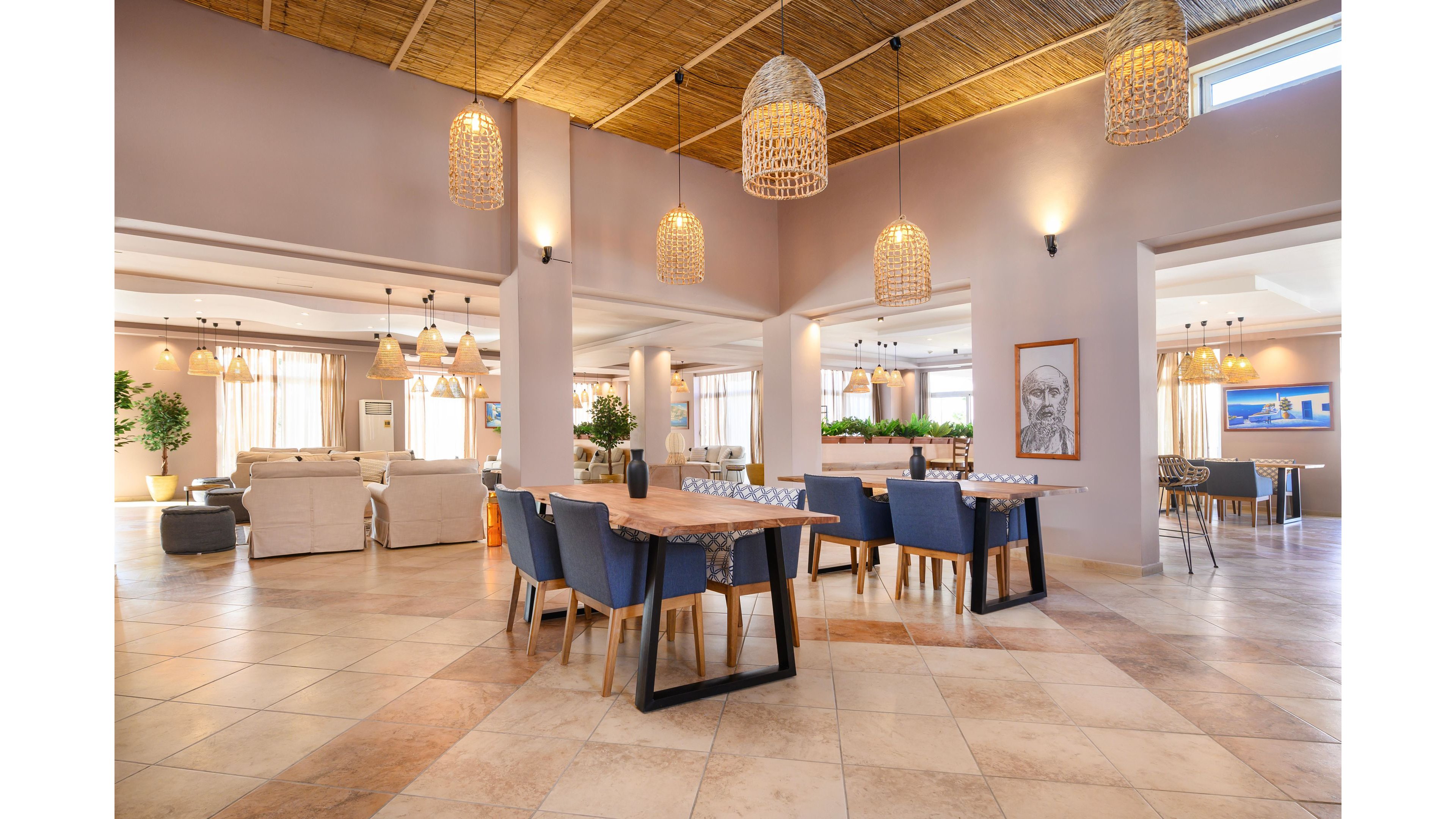 Spacious lobby lounge with modern tables and blue upholstered chairs is shown in warm natural light. Large woven pendant lamps hang from a wooden ceiling above tiled floors, creating a relaxed, airy atmosphere. Sofas and armchairs form seating areas in the background, with potted plants adding greenery throughout the space. Wall art and wide windows with sheer curtains contribute to the contemporary, welcoming design.