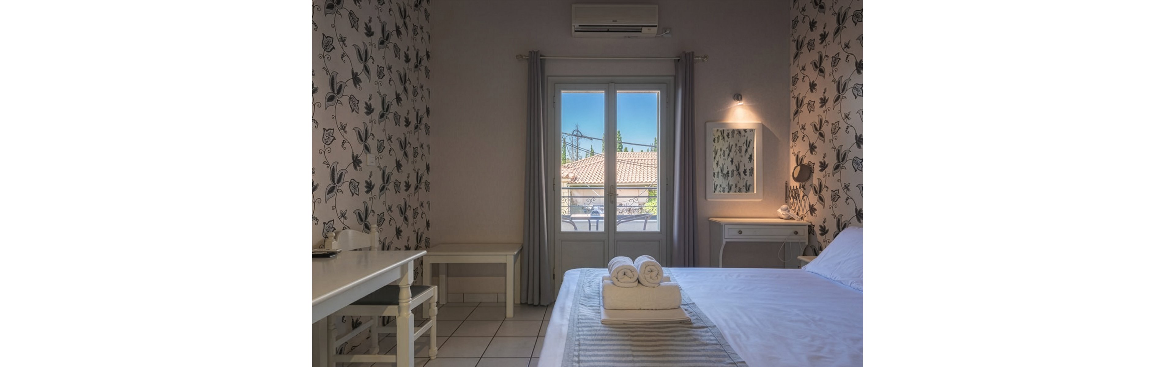 A neatly arranged bedroom features a large bed with white linens and rolled towels placed on top. The room includes a small desk, chair, and side table, all in light tones that complement the floral patterned wallpaper. Sunlight filters through glass doors leading to a balcony, revealing tiled rooftops and trees outside. Soft lighting and neutral colors create a calm, inviting atmosphere suitable for rest or vacation stays.