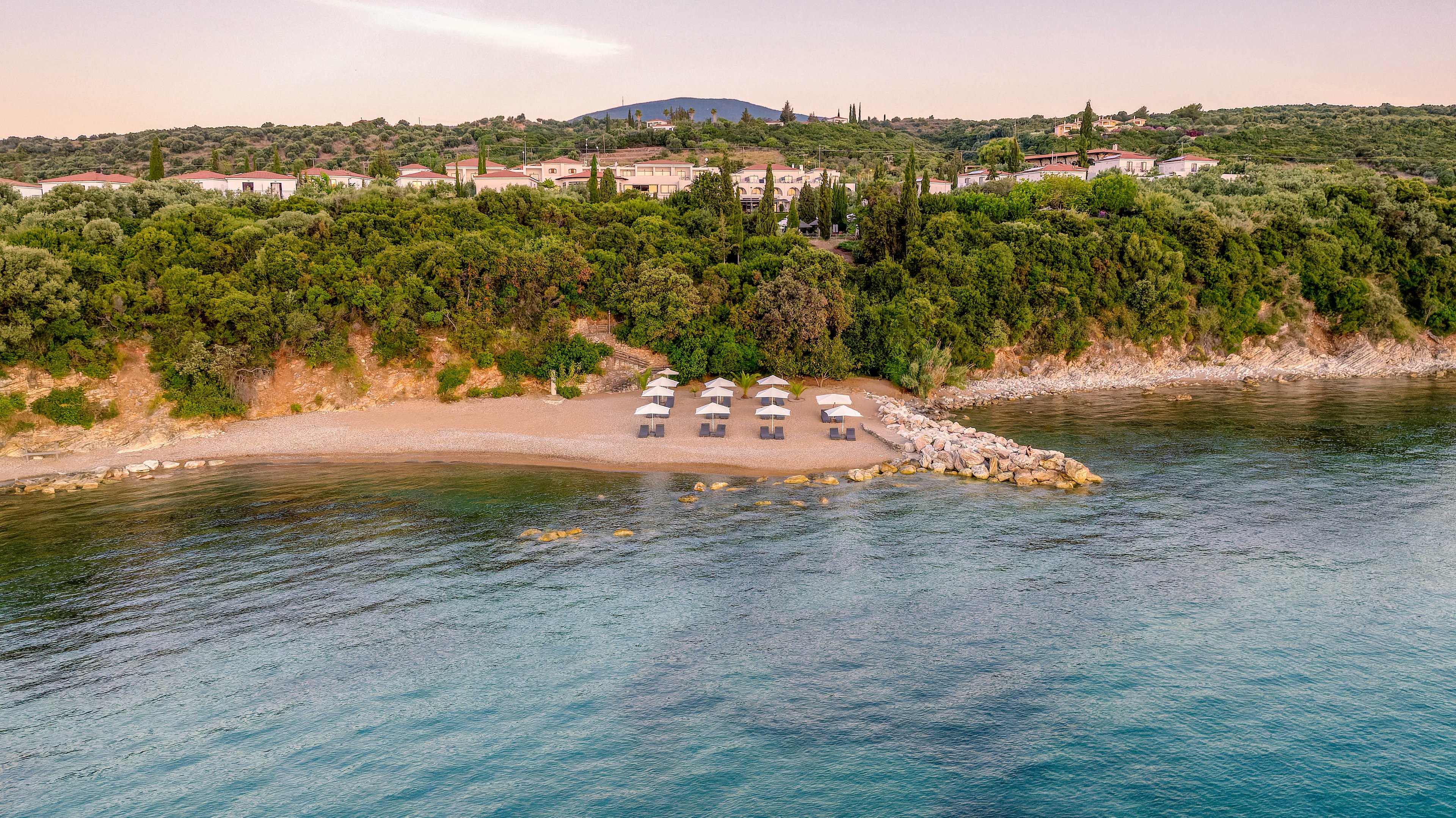 An aerial view shows a small secluded beach lined with neatly arranged sun loungers and umbrellas. The beach is bordered by calm, clear water in the foreground and a dense green forest on the hillside behind. Buildings are partially visible above the tree line, suggesting a coastal resort or village. The soft evening light creates a tranquil, inviting atmosphere along the shoreline.