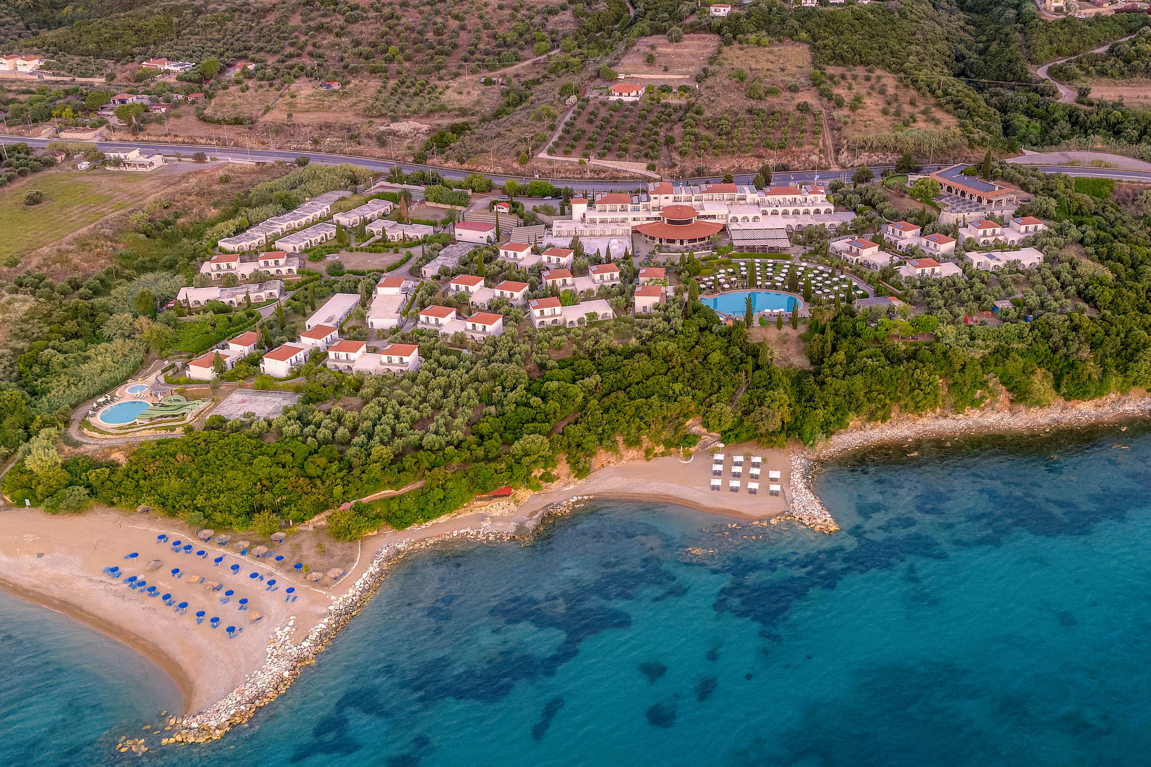 A large seaside resort complex is captured from above, nestled between lush greenery and a calm turquoise sea. Rows of white buildings with red roofs are arranged around gardens, pools, and pathways. The sandy beach in the foreground features neatly organized sun loungers and umbrellas along the shoreline. Beyond the resort, a road and terraced hills with scattered vegetation frame the scene, emphasizing the remote coastal setting.
