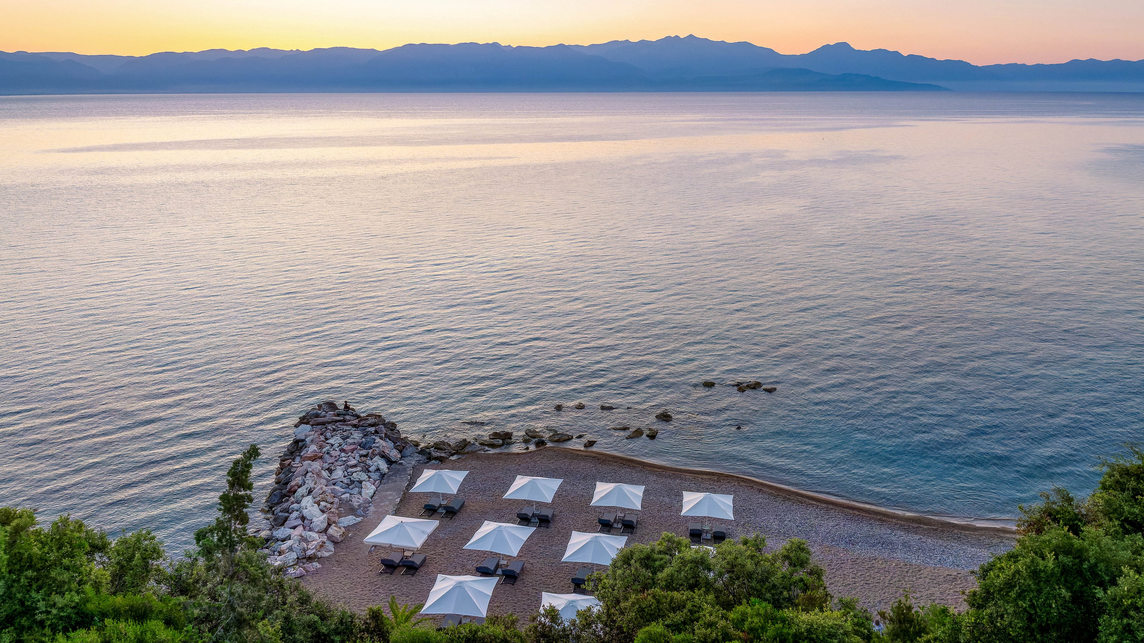 A quiet pebble beach resort is shown at sunrise, with several white cabanas and lounge chairs neatly arranged along the shoreline. Calm sea water stretches into the distance, reflecting the soft pastel colors of the early morning sky. A rocky jetty extends into the water on the left, while lush green vegetation frames the foreground. Distant mountains create a serene backdrop, emphasizing the peaceful coastal atmosphere.