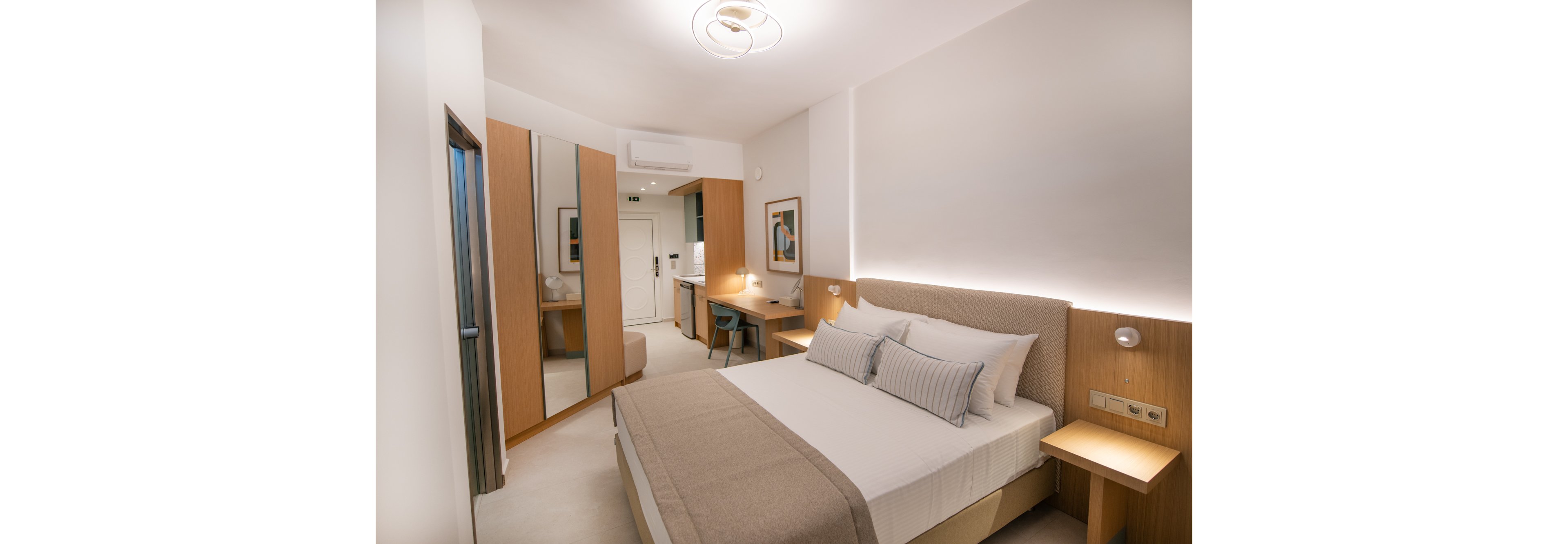 A contemporary hotel room features a neatly made double bed with neutral bedding and accent pillows. The space includes a built-in desk, chair, and wall-mounted lighting, with a large mirror and minimalist artwork adding visual interest. Warm wood tones and soft lighting create a welcoming, tranquil atmosphere. The room is uncluttered, with a clean, modern aesthetic and no visible people or text.