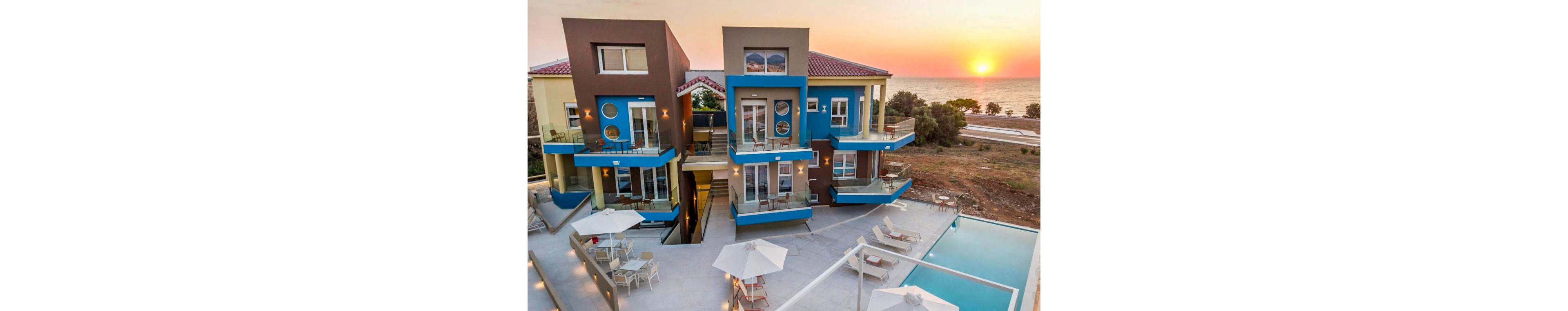 A contemporary hotel complex features geometric architecture with blue accents, situated by the sea. The outdoor area includes a swimming pool, sun loungers, and shaded tables, all overlooking a scenic sunset. The setting is tranquil, with minimal activity and a warm, inviting atmosphere. The ocean and horizon are visible in the background, enhancing the vacation vibe. No visible text or numbers are present in the image.
