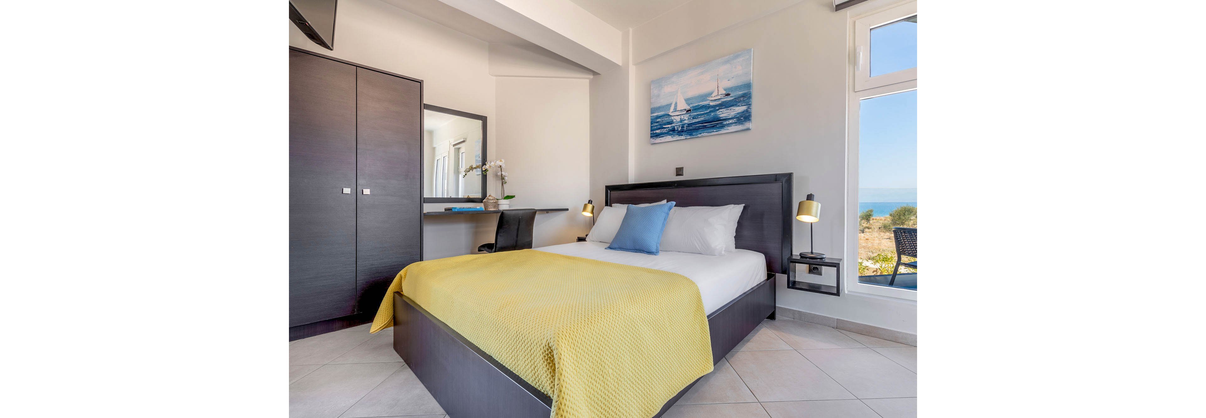 A contemporary hotel room features a double bed with a yellow blanket and blue accent pillow. The space is bright, with large windows offering a scenic view of the sea and landscape outside. Minimalist decor includes a painting of sailboats, a dark wardrobe, and a desk with a mirror and orchid. The room is well-lit with bedside lamps and natural light, creating a fresh and inviting atmosphere.