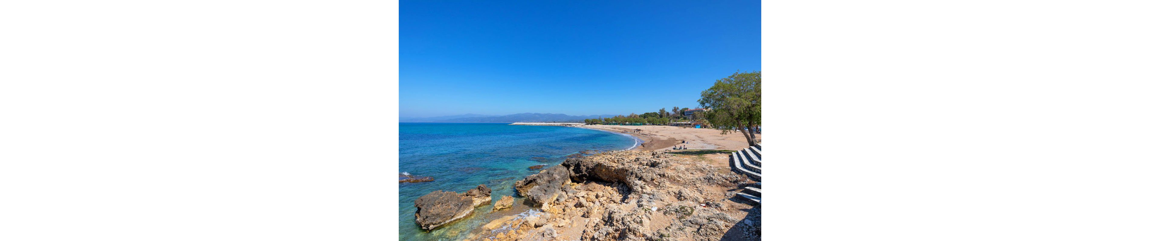 A tranquil beach scene features rocky formations in the foreground and a sandy shoreline curving into the distance. The clear blue sea meets a bright sky, with a few people visible enjoying the peaceful setting. Trees provide shade along the edge, and distant mountains are faintly visible. The overall mood is calm and inviting, with natural colors dominating the palette.
