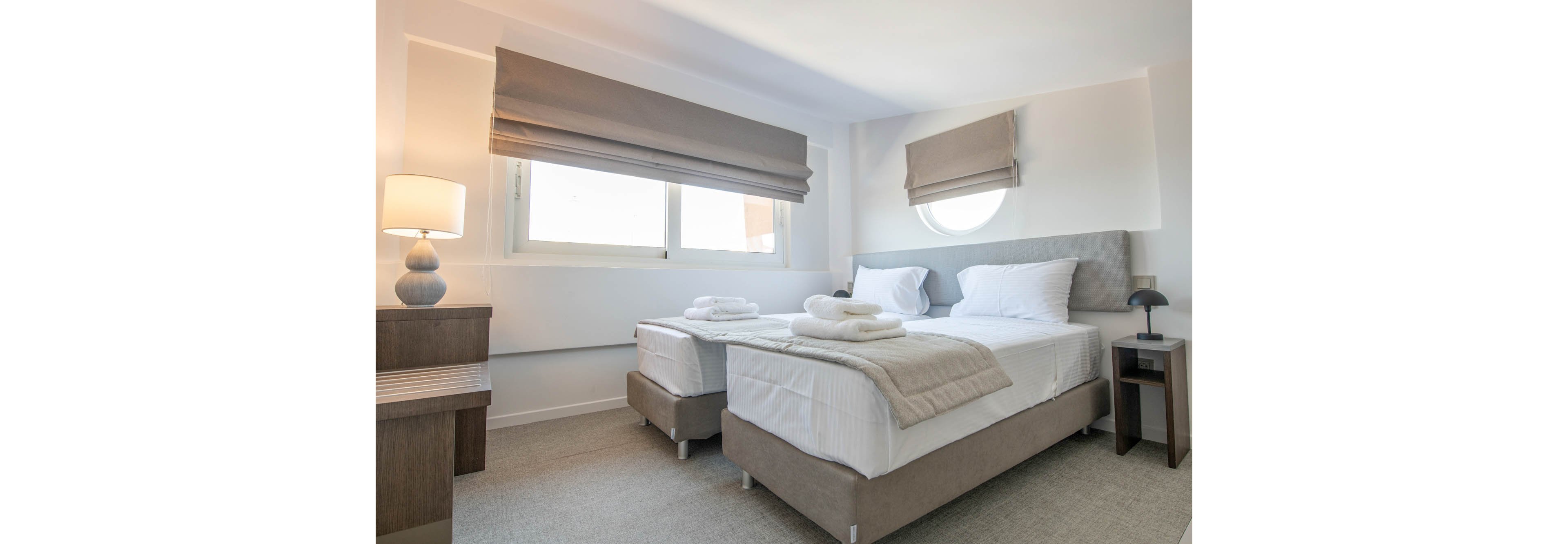 A contemporary hotel room features two neatly made twin beds with white linens and folded towels on top. The space is illuminated by natural light from large windows with taupe roman shades, complemented by a soft-glow table lamp. The decor is minimalist with neutral tones, creating a calm and inviting atmosphere. No visible text or numbers are present in the image.