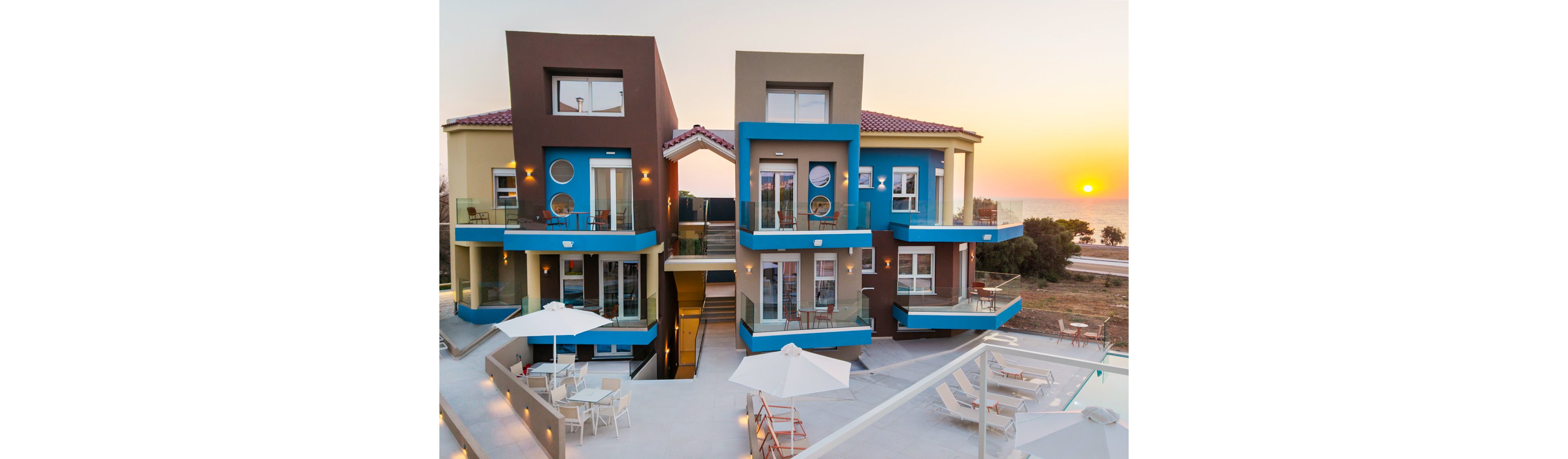 Contemporary apartment buildings with bold blue and brown accents overlook a serene beach at sunset. The setting features a spacious outdoor terrace with white umbrellas, tables, and a swimming pool in the foreground. The architecture is geometric and modern, with large windows and balconies facing the sea. The sun is setting over the horizon, casting a warm glow across the scene.