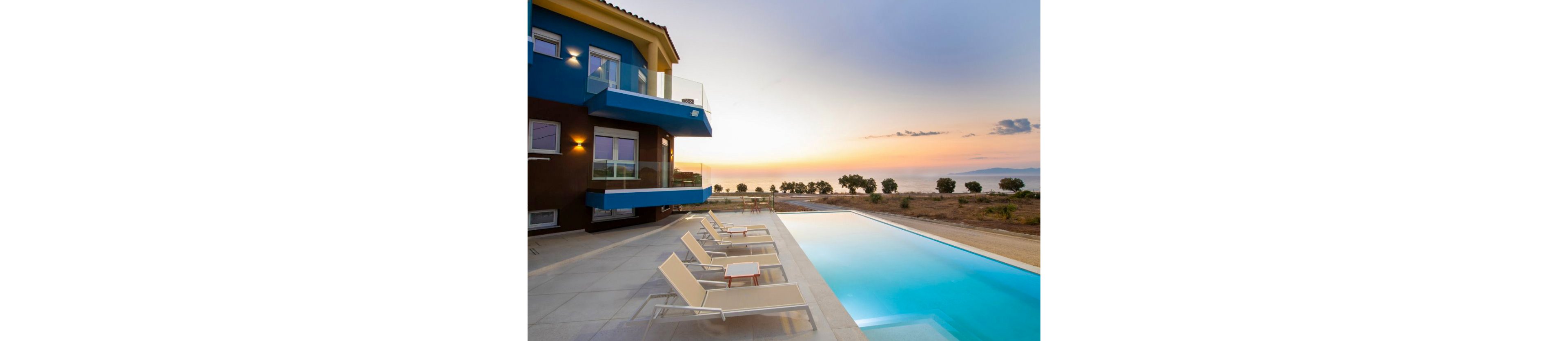 A contemporary villa features a sleek outdoor swimming pool with several lounge chairs arranged on the poolside patio. The setting captures a tranquil sunset, with soft pastel colors in the sky and a scenic view of the sea and distant trees. The building’s modern architecture is highlighted by clean lines and blue accents, creating a relaxing and inviting atmosphere. No visible text or numbers are present in the image.