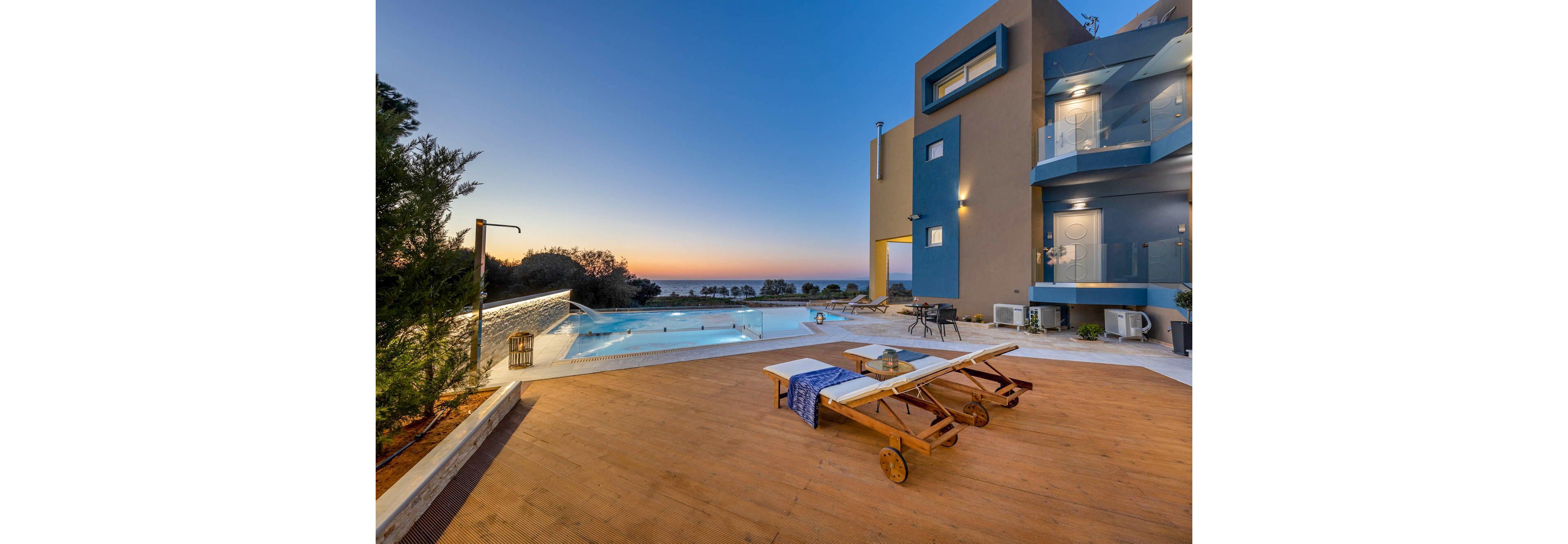 A contemporary villa features a spacious outdoor pool area overlooking the ocean at sunset. Two wooden lounge chairs with towels and drinks are set on a wooden deck, creating a relaxing atmosphere. The building's modern architecture is highlighted by glass balconies and soft exterior lighting, while the tranquil setting enhances the luxurious mood.