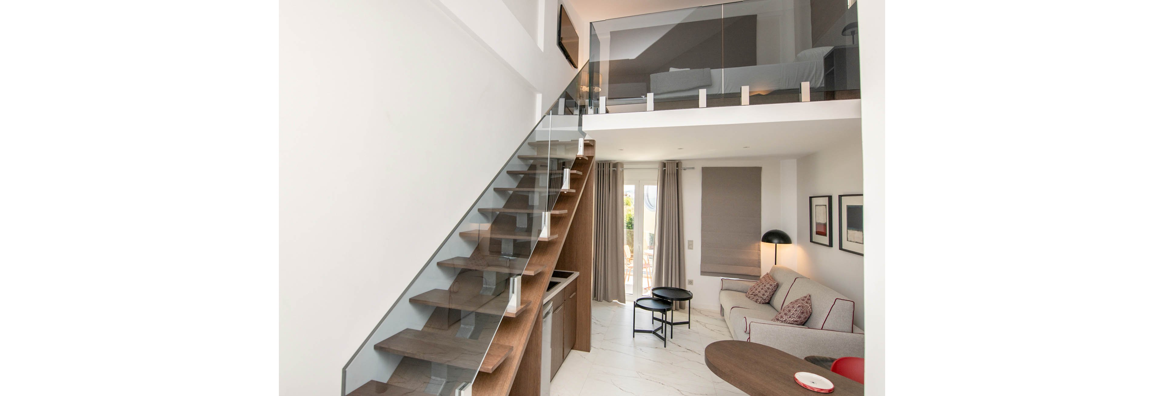 A contemporary loft apartment features a sleek glass staircase leading to an upper mezzanine. The main living area includes a sofa, round coffee tables, and framed wall art, with large windows letting in natural light. The interior palette is neutral with accents of wood and black, creating a clean and inviting atmosphere. No visible numeric values or identifiable brands are present.