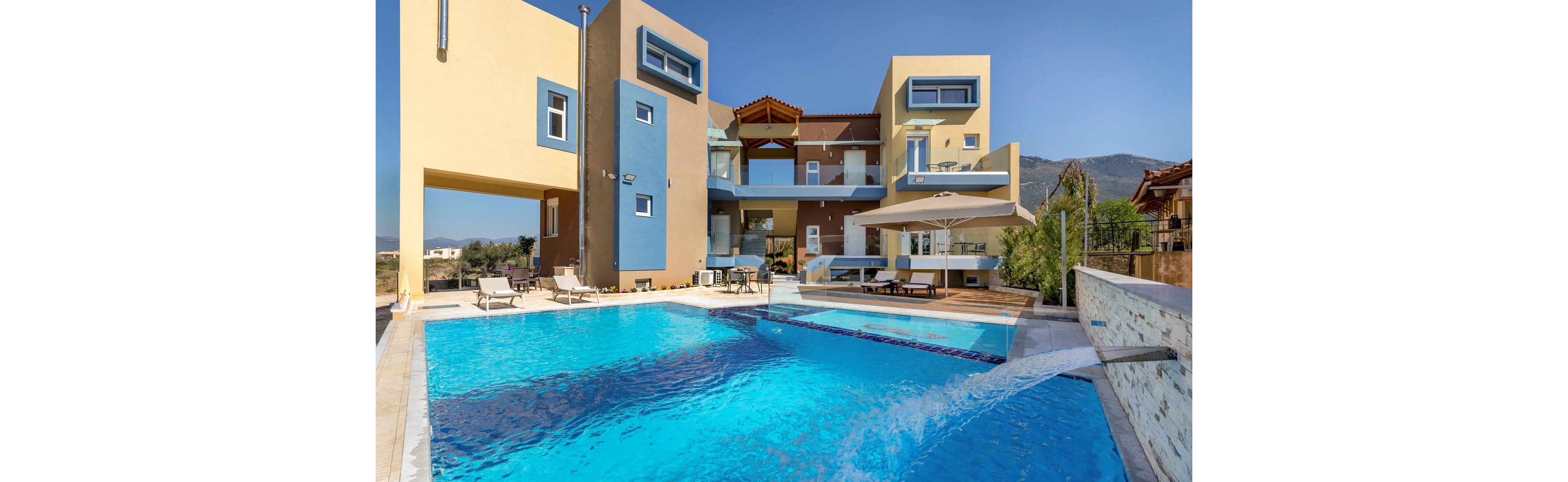 A contemporary multi-story villa features bold geometric architecture and a vibrant color palette. The scene is set outdoors with a large, clear blue swimming pool in the foreground, surrounded by sun loungers and a shaded patio area. The building's clean lines and open balconies create a relaxed, luxurious atmosphere under a bright, sunny sky. No visible text or numbers are present in the image.