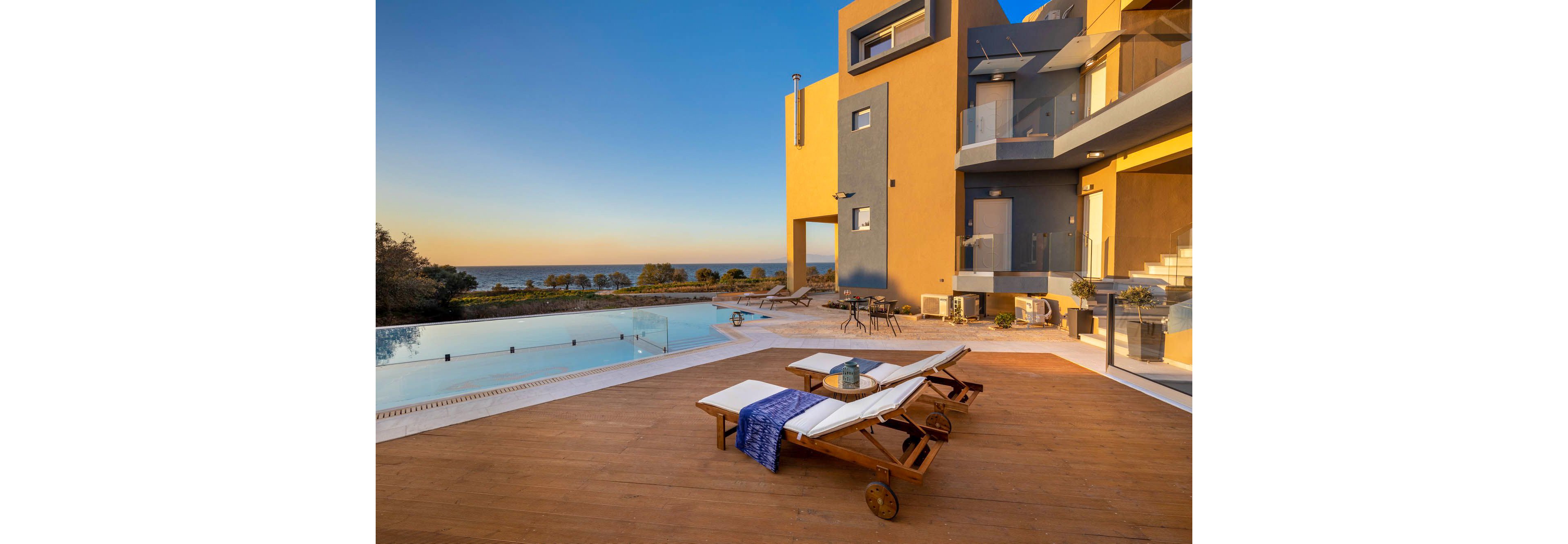 A contemporary villa overlooks the ocean, featuring a spacious wooden deck with two cushioned lounge chairs and a small table. The setting includes an inviting infinity pool and modern architectural elements in warm tones. The scene is bathed in golden sunset light, creating a tranquil and luxurious atmosphere. No visible text or numbers are present in the image.