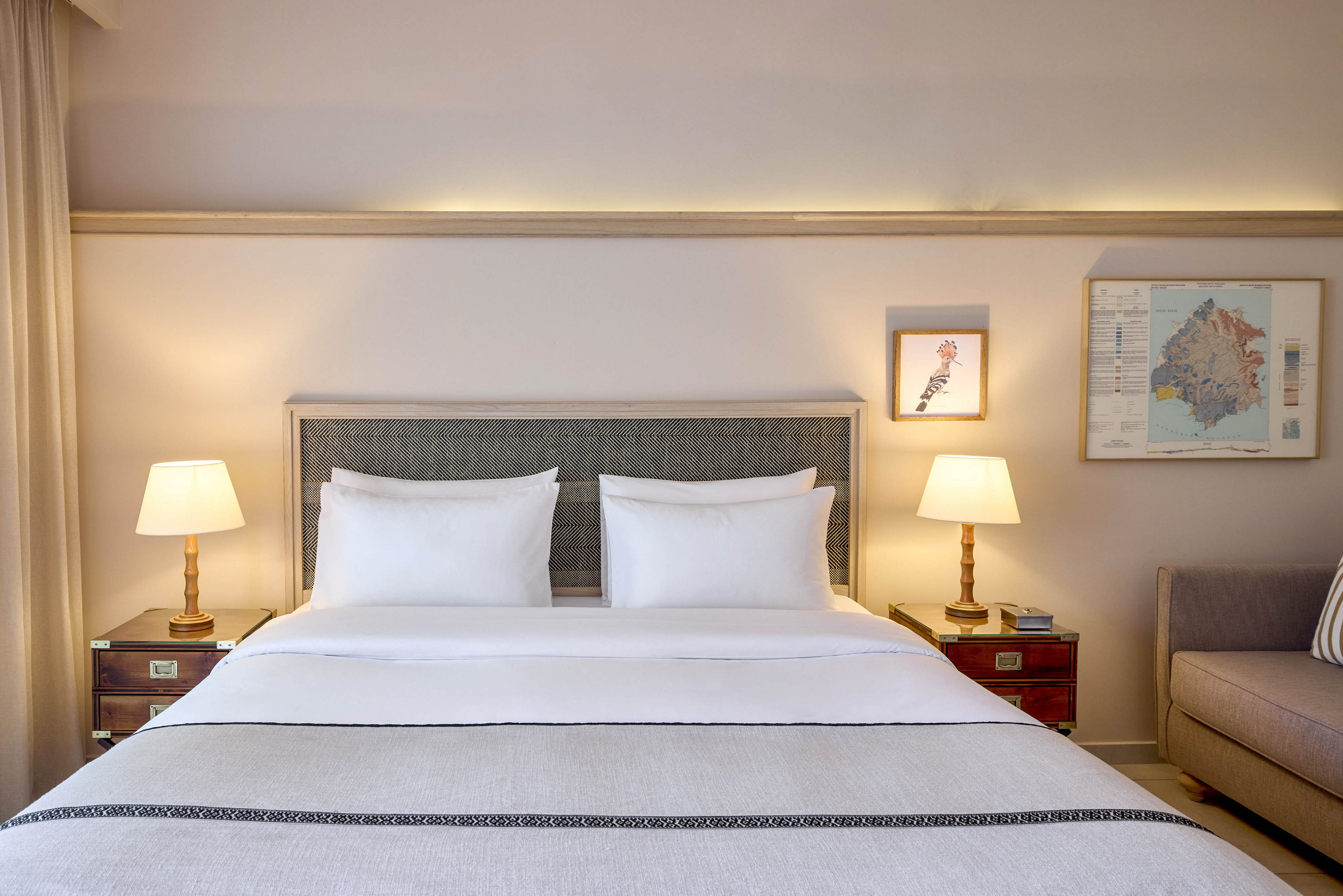A neatly made double bed with white linens is centered in a softly lit hotel bedroom. Two bedside tables with matching lamps flank the bed, casting a warm glow on the neutral walls. Framed artwork hangs above the headboard, and a sofa is visible to the side, enhancing the room’s comfort. The overall design is minimalist and inviting, with a calm, contemporary style.