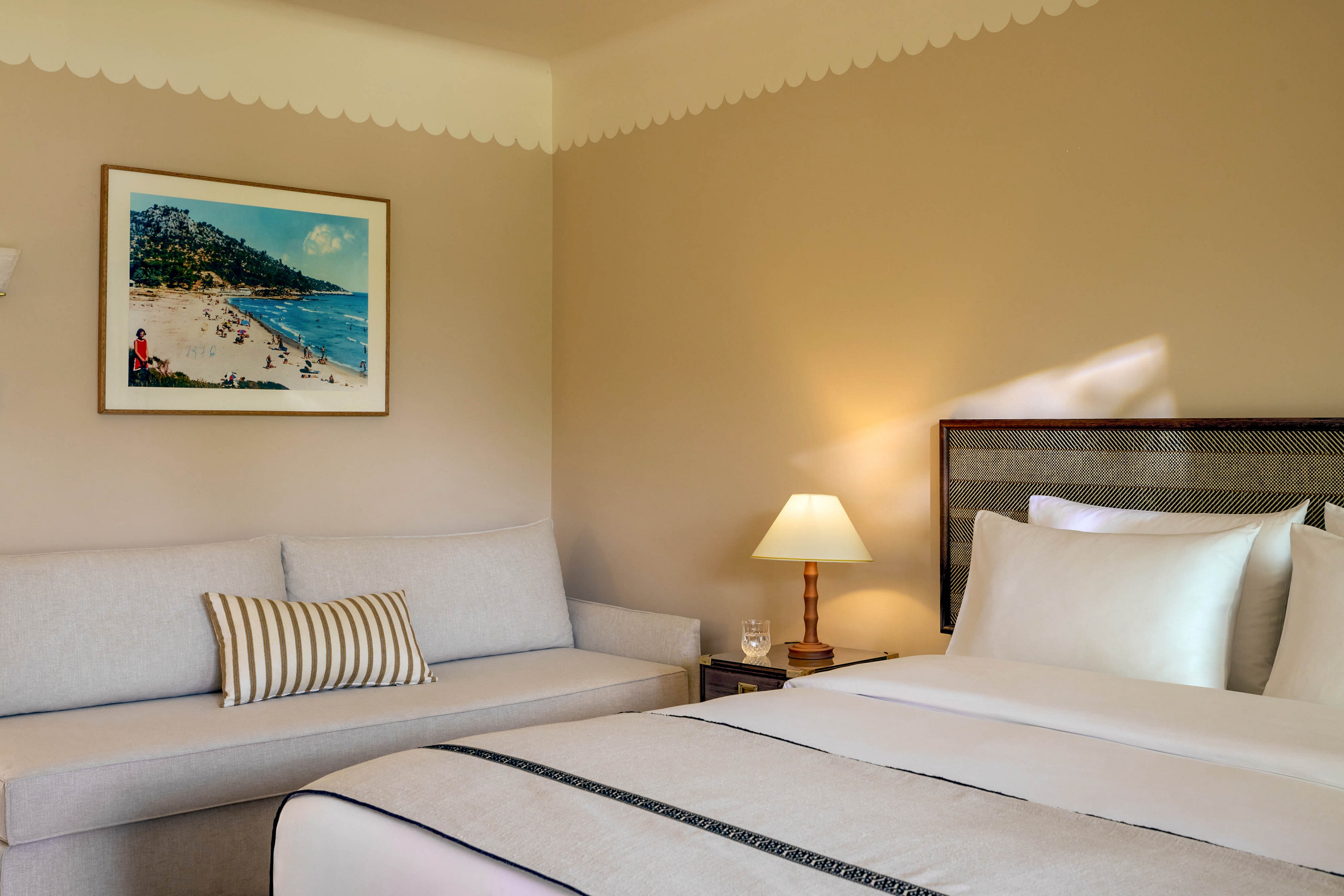 A neatly made bed with crisp white linens is the focal point of this cozy hotel bedroom. Beside the bed, a cushioned sofa with a striped pillow provides additional seating. A bedside table holds a lit lamp and a glass of water, adding warmth and comfort to the scene. A framed beach painting hangs on the wall, bringing a touch of coastal charm to the neutral-toned room.