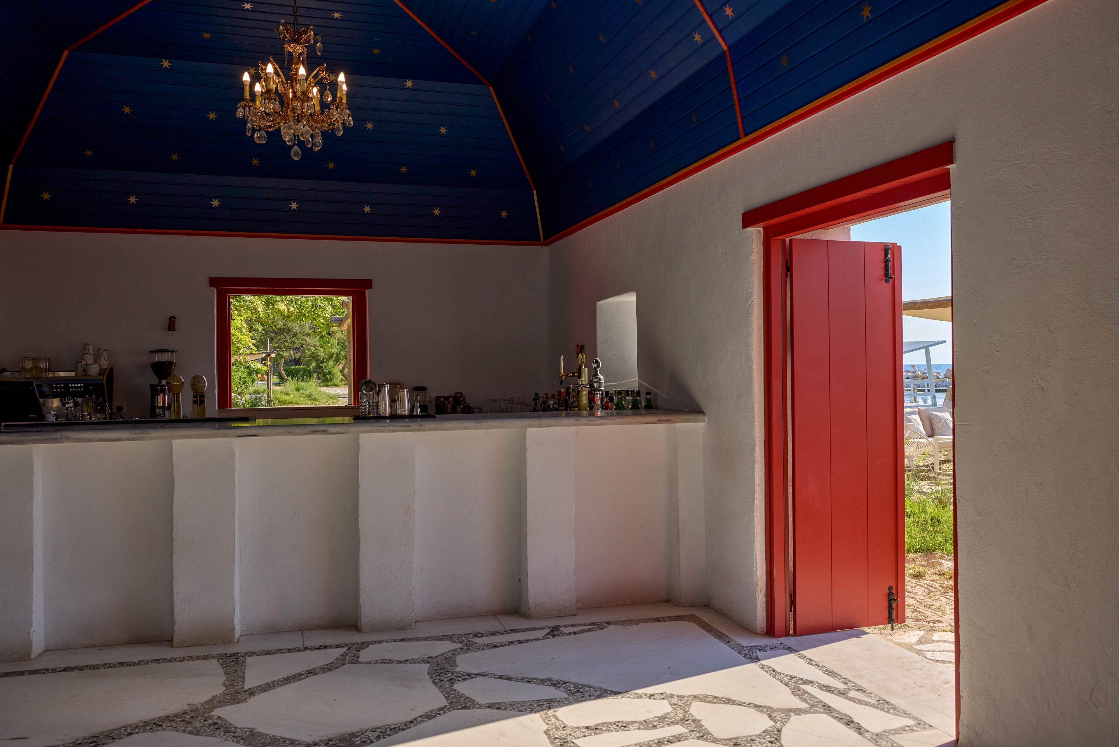 An intimate bar interior features a long white counter and a small window looking out to greenery. The space has a striking blue vaulted ceiling with decorative golden dots and a classic chandelier. Bright red trim around the door and window contrasts with the neutral walls and stone-patterned floor. Sunlight streams through the open door, revealing an outdoor seating area near the water.