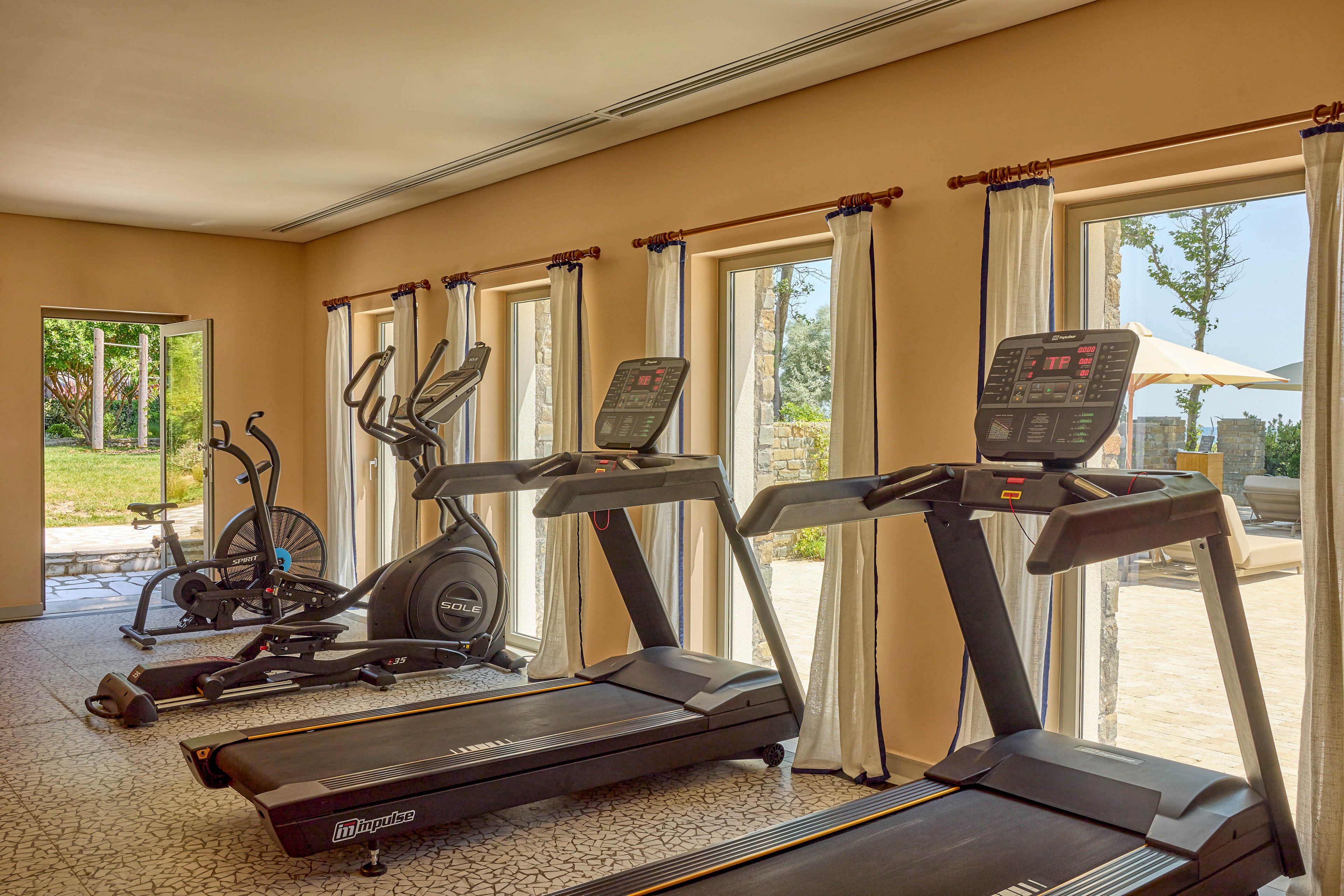 A sunlit indoor gym features multiple treadmills and stationary bikes arranged in a row. Large floor-to-ceiling windows with light curtains open onto a green outdoor garden and patio area. The neutral beige walls and patterned floor tiles create a calm, inviting workout environment. Exercise equipment displays are visible, adding a modern, professional feel to the space.