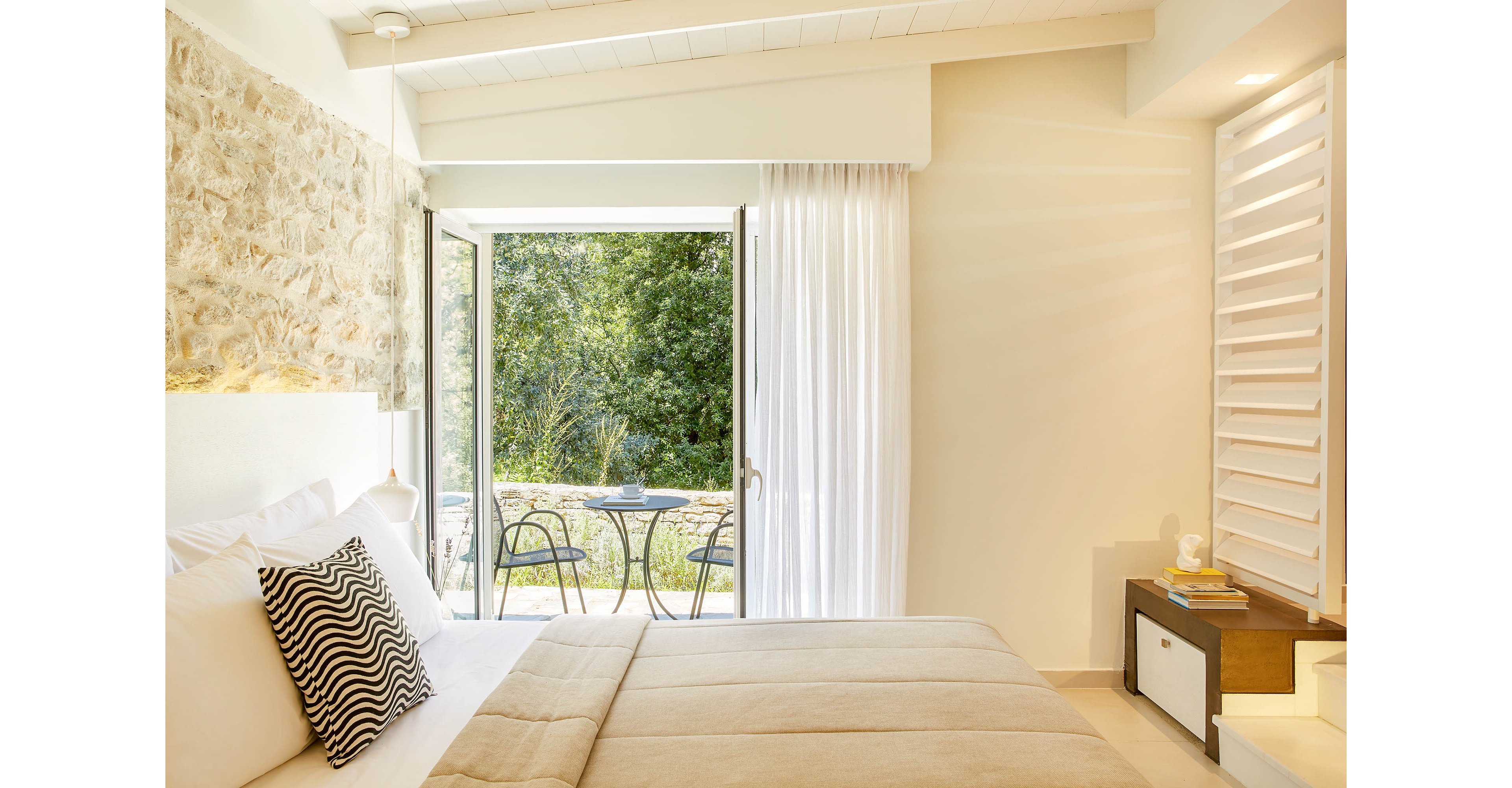 A cozy, modern bedroom features a neatly made bed with neutral bedding and a patterned accent pillow. Large glass doors open onto a small patio with two metal chairs and a round table, overlooking lush greenery. Natural light floods the room, highlighting the stone accent wall and clean white ceiling. A bedside table with books and a lamp adds a warm, inviting touch to the serene space.