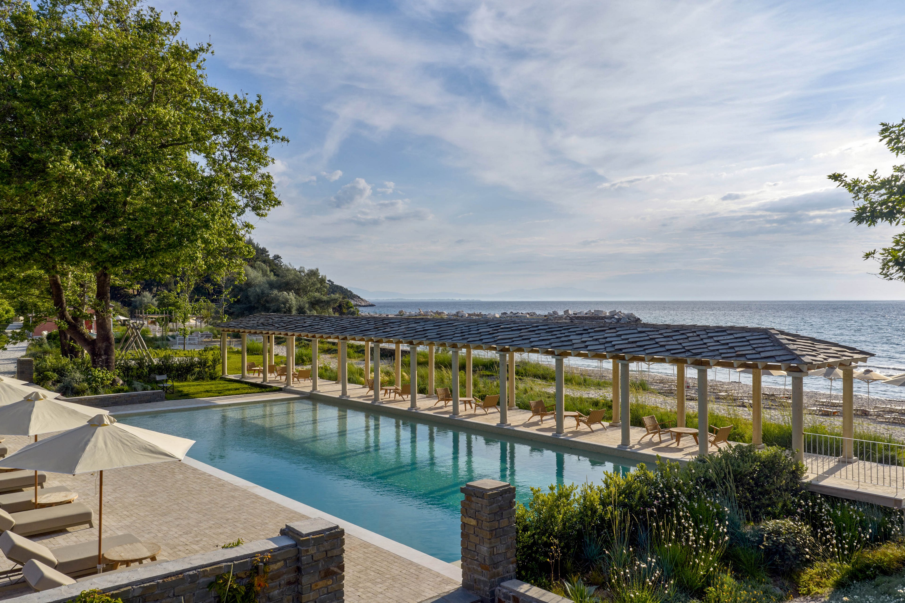 An elegant outdoor swimming pool stretches alongside a sandy shoreline at a seaside resort. A long pergola with lounge chairs runs parallel to the water, offering shaded relaxation with views of the ocean. Lush greenery, trees, and landscaped gardens frame the pool area, creating a tranquil atmosphere. Soft morning or late afternoon light enhances the calm, vacation-like mood of the scene.