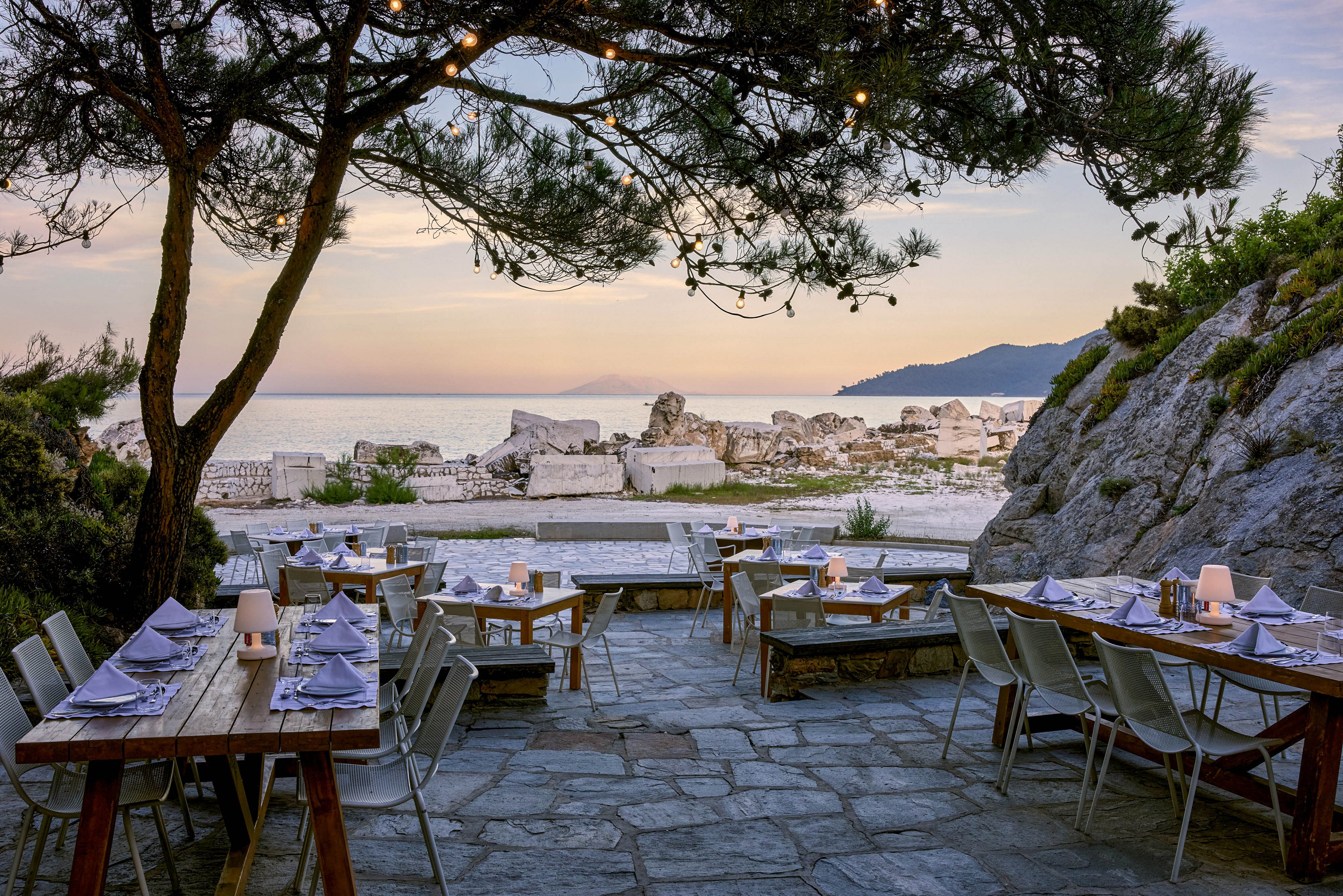 An outdoor seaside restaurant is set with wooden tables and neatly arranged place settings under a large tree. The dining area overlooks a calm sea and distant mountains at sunset, creating a tranquil coastal atmosphere. String lights hang above the stone patio, adding warmth to the scene. Empty chairs and folded napkins suggest the space is prepared for evening guests in a relaxed yet elegant setting.