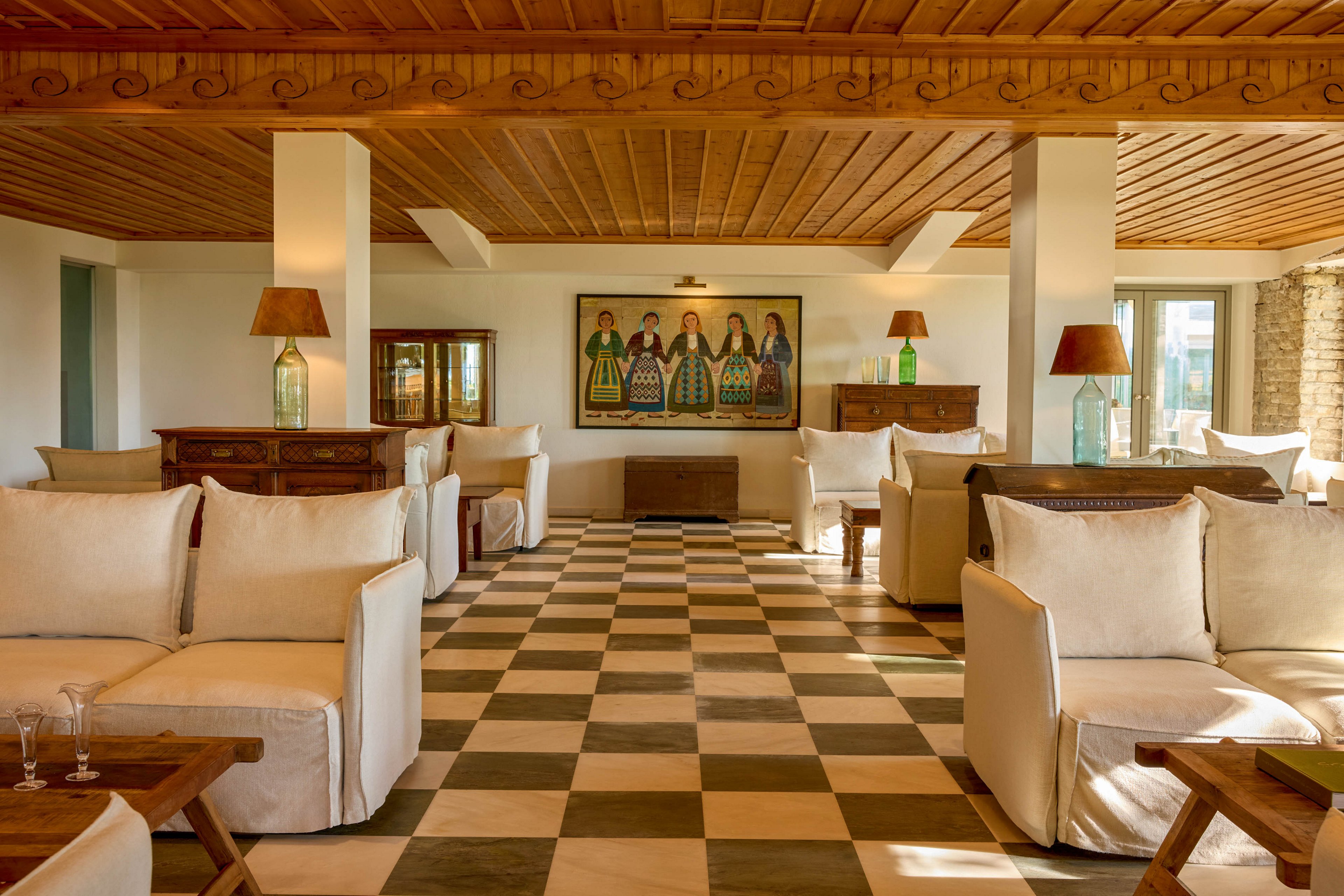 A warm, sunlit lounge features plush white sofas arranged around low wooden tables. The room has a distinctive checkered floor in neutral tones and a wooden plank ceiling with decorative trim. Tall table lamps with brown shades and glass bases flank rustic wooden chests along the walls. A colorful framed artwork depicting several figures hangs prominently on the far wall, adding a focal point to the cozy interior.