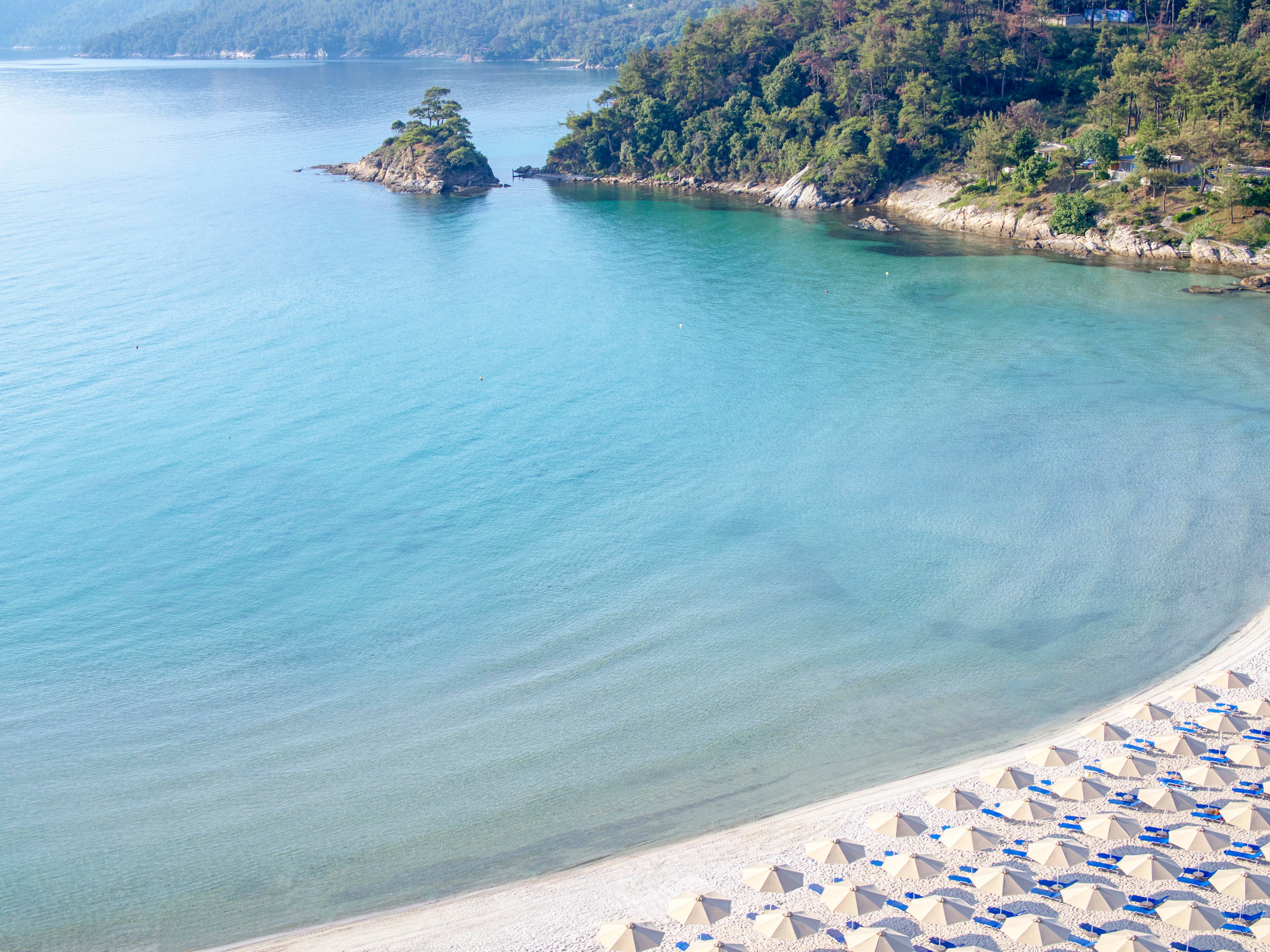 A serene coastal bay with calm turquoise water curves gently along a sandy shoreline. Neatly arranged rows of closed beach umbrellas and sun loungers line the empty beach in the foreground. A small tree-covered islet and forested hills frame the background, reflecting softly on the water. The scene suggests a peaceful, early morning or off-season seaside resort atmosphere.