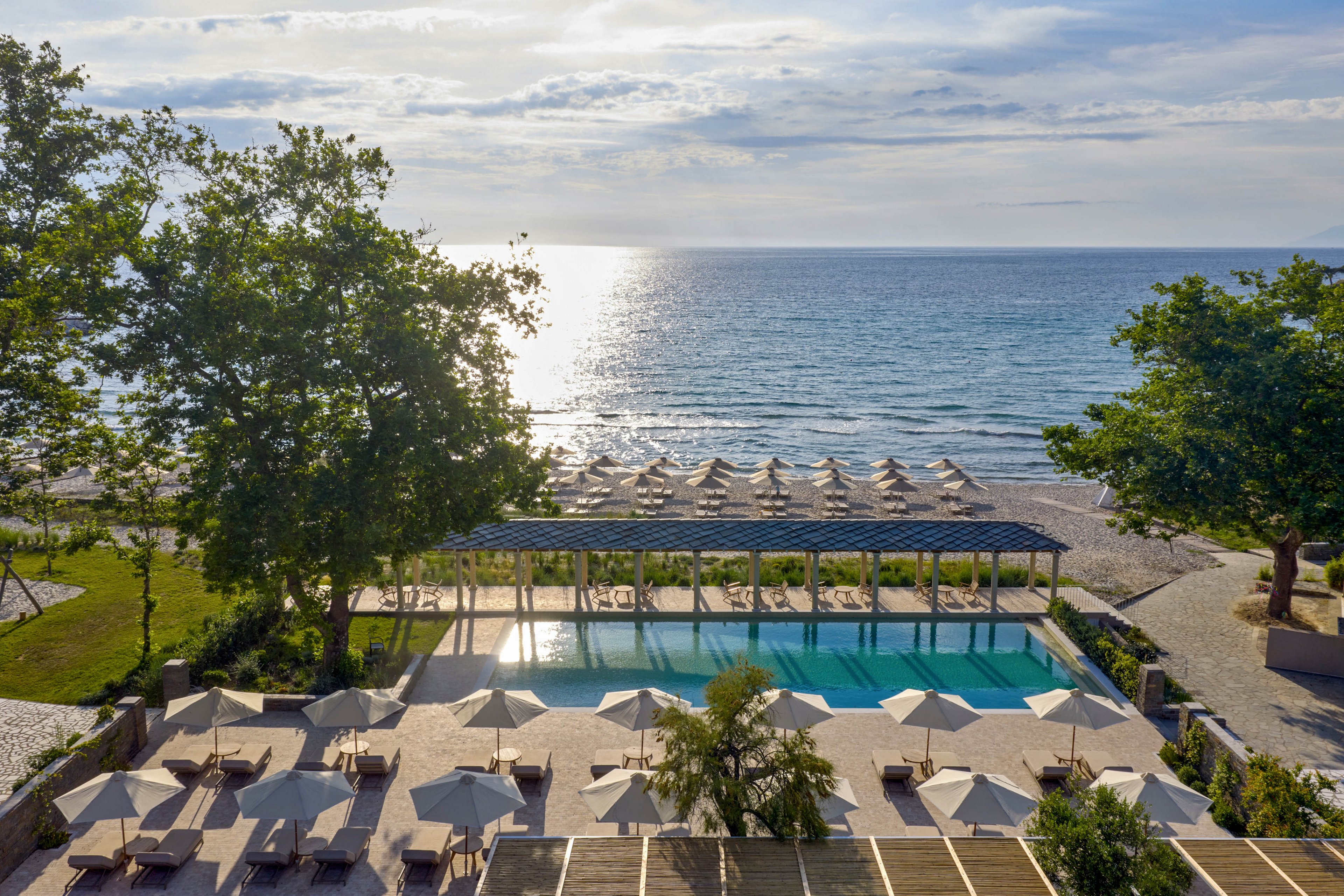 An elevated view shows a resort swimming pool overlooking a calm sea. Numerous sun loungers and umbrellas are arranged neatly around the pool and along the sandy beach. Tall green trees frame the scene, adding shade and contrast to the bright paved terraces. The sky is partly cloudy, creating a serene and inviting coastal atmosphere.