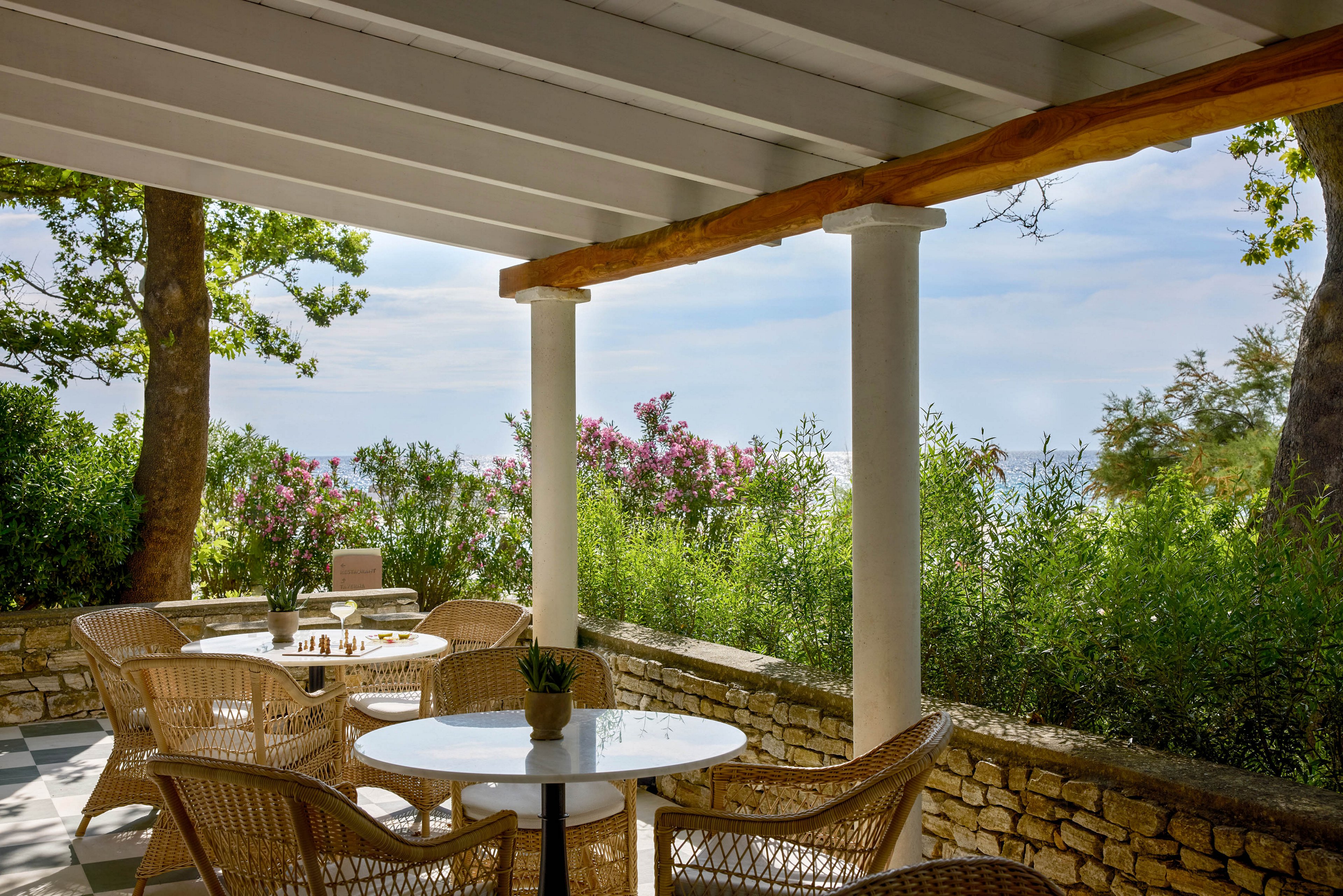 An inviting outdoor terrace features round white tables and wicker chairs under a shaded pergola. The seating area overlooks dense green shrubs and flowering plants, with glimpses of the sky and distant landscape beyond. Soft natural light filters through the trees, creating a calm and relaxing atmosphere. A small plant centerpiece and a chessboard on the tables add casual, leisurely details to the scene.