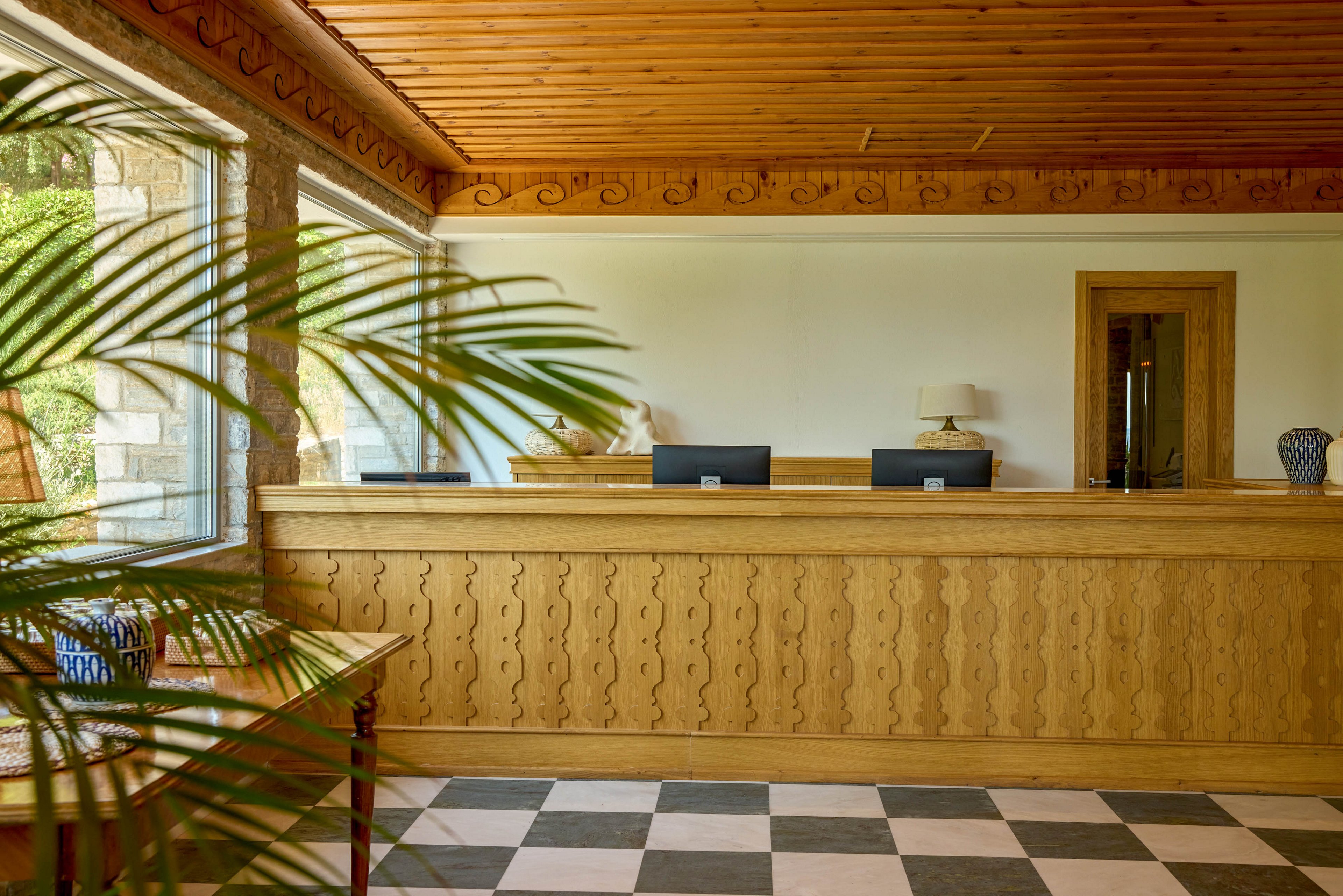 A bright hotel reception area features a long wooden front desk with carved detailing. Two computer monitors sit on the counter, accompanied by a table lamp and decorative vase. Large windows on the left let in natural light and reveal greenery outside. The space is finished with a wooden ceiling, white walls, and a black-and-white checkered floor, creating a warm yet clean atmosphere.