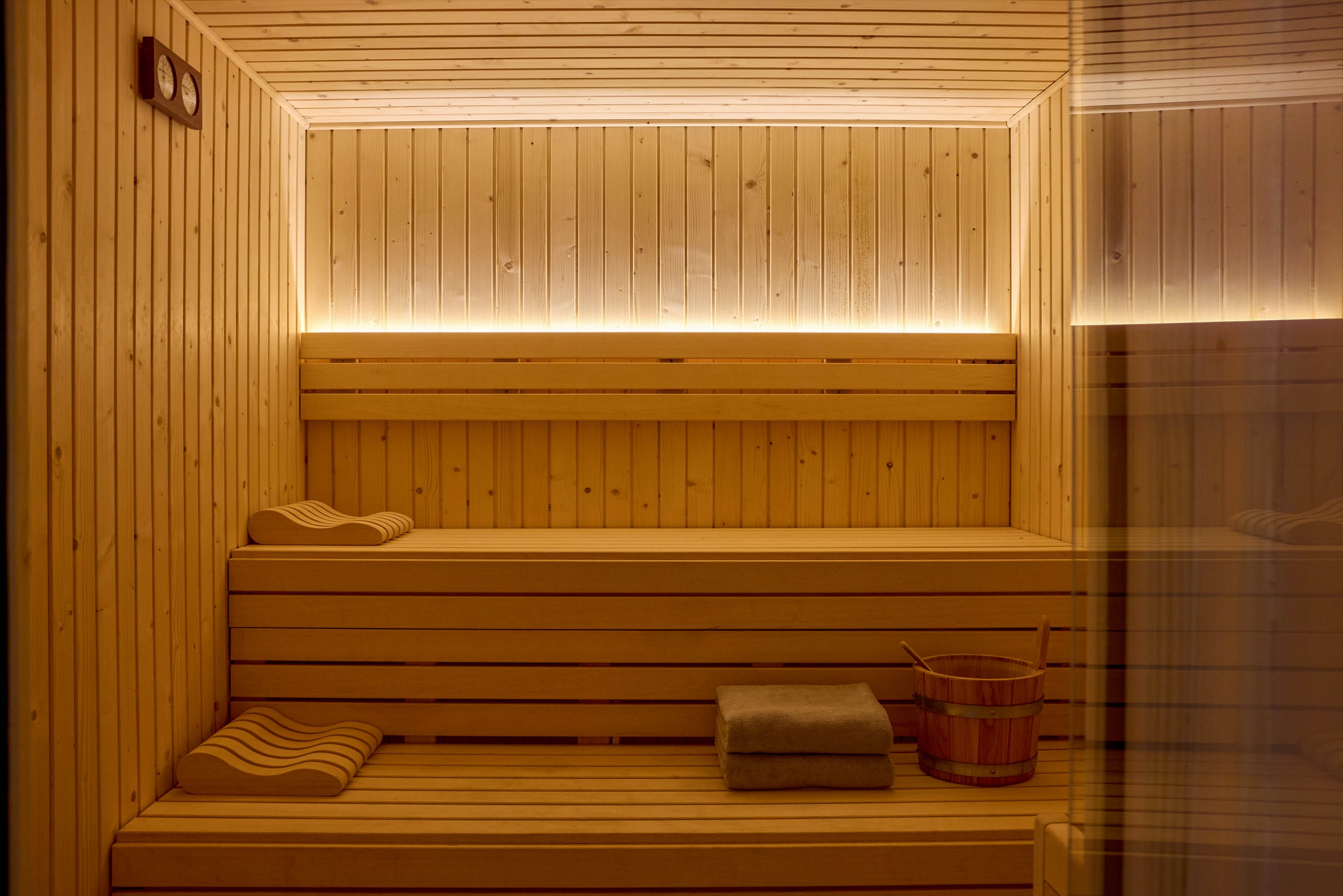 A cozy wooden sauna interior features tiered benches made of light-colored timber. Soft indirect lighting runs along the walls, creating a warm and inviting glow. A folded towel and a wooden bucket with a ladle rest on the lower bench, suggesting a relaxing spa experience. The clean lines and natural materials emphasize a minimalist, tranquil atmosphere.
