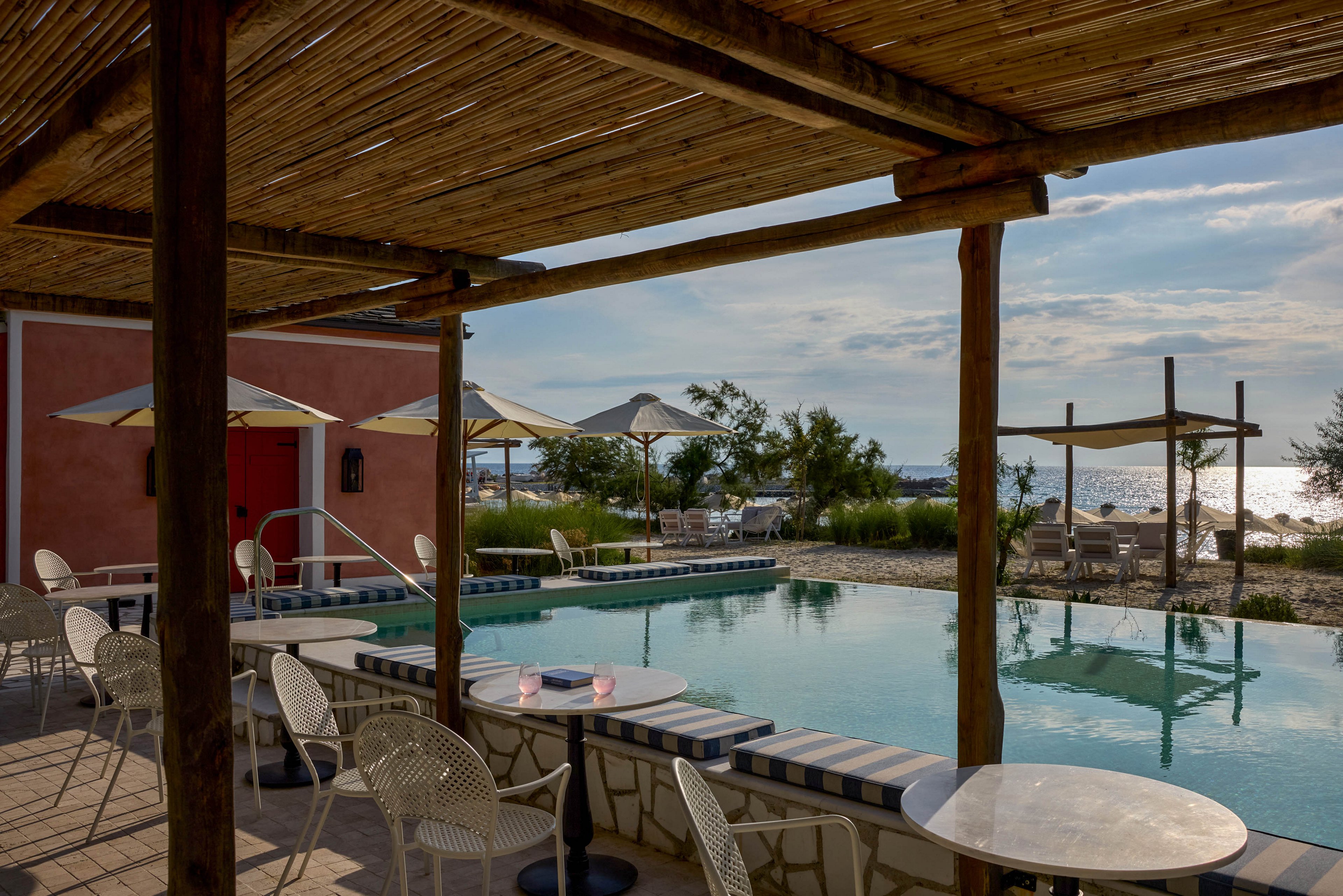 An outdoor café terrace overlooks a small swimming pool near the sea. White chairs and round tables sit beneath a wooden pergola, with cushioned seating lining the pool edge. Large umbrellas and sparse greenery frame the view toward the shoreline and calm water. Soft afternoon light and partly cloudy skies create a relaxed coastal atmosphere.