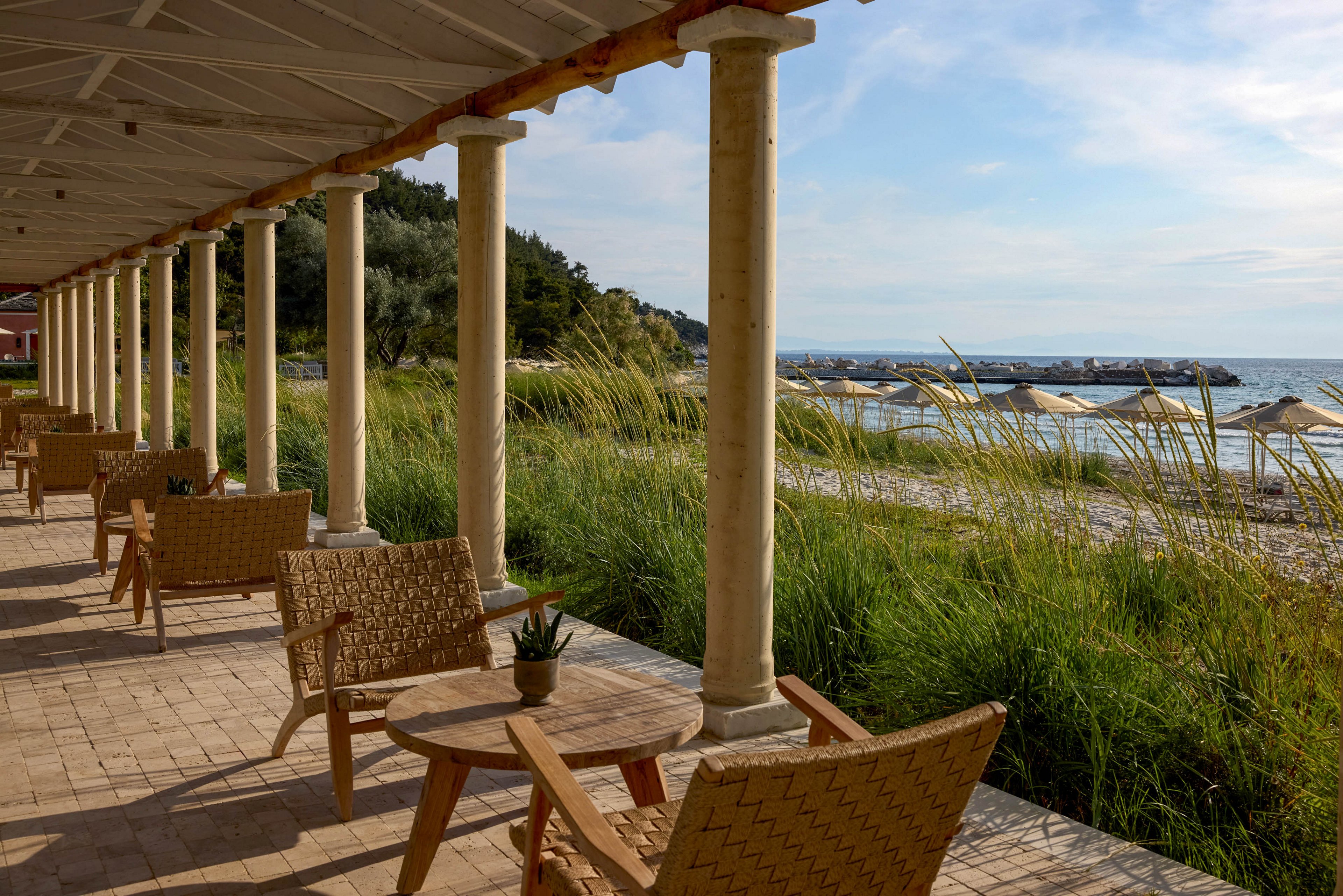 An open-air seaside terrace features wicker chairs and round wooden tables arranged in a row beneath a covered colonnade. The view looks out toward a sandy beach bordered by tall grasses and calm water. Sun umbrellas and distant rocks line the shoreline under a partly cloudy sky. Warm light and natural textures create a relaxed, coastal atmosphere ideal for outdoor lounging.