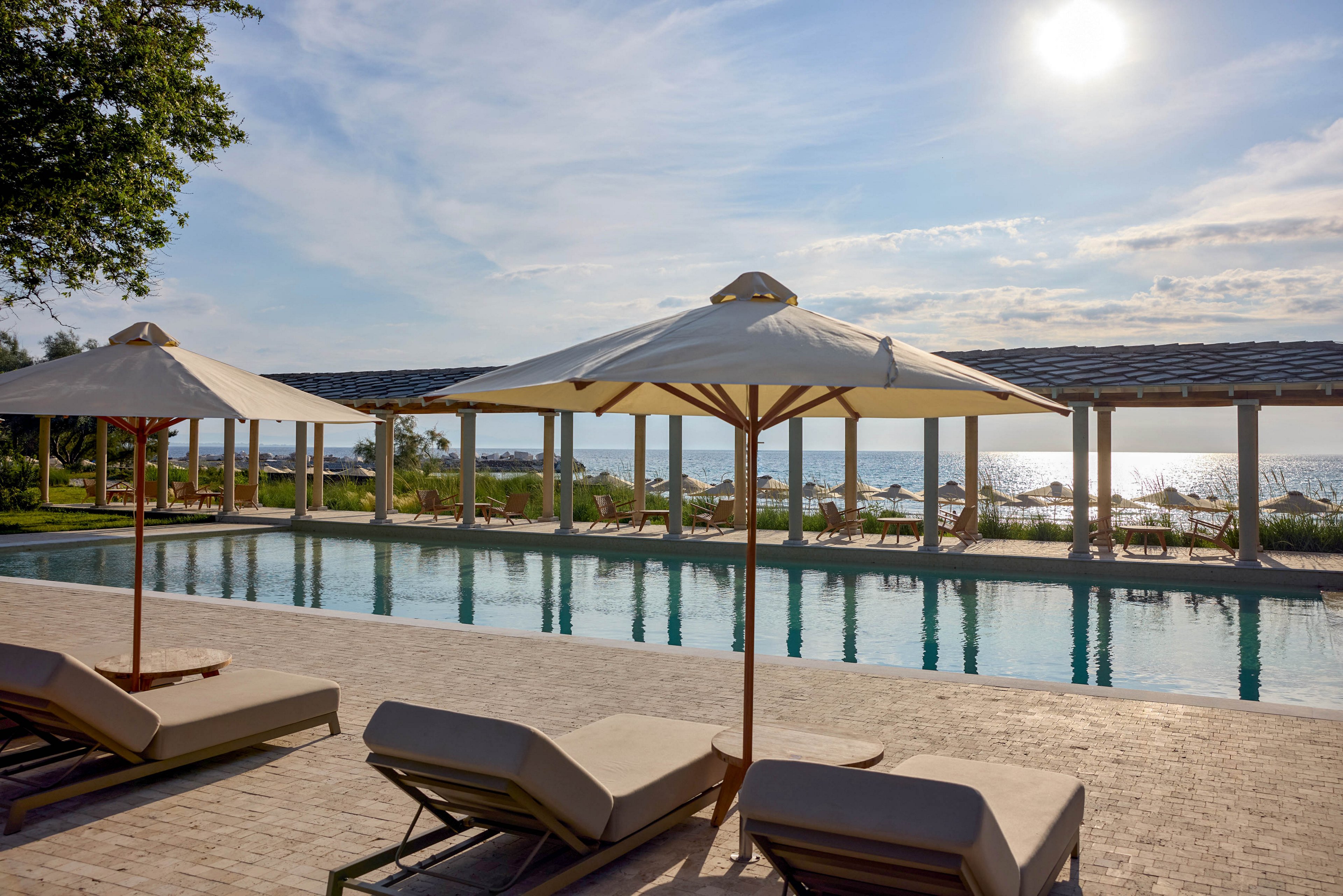 An outdoor resort-style swimming pool is shown under a bright sunny sky. Lounge chairs with cushions and large umbrellas line the poolside terrace, creating a relaxing atmosphere. In the background, a covered walkway and distant water view suggest a coastal or beachfront setting. The scene feels calm and inviting, ideal for leisure and vacation relaxation.