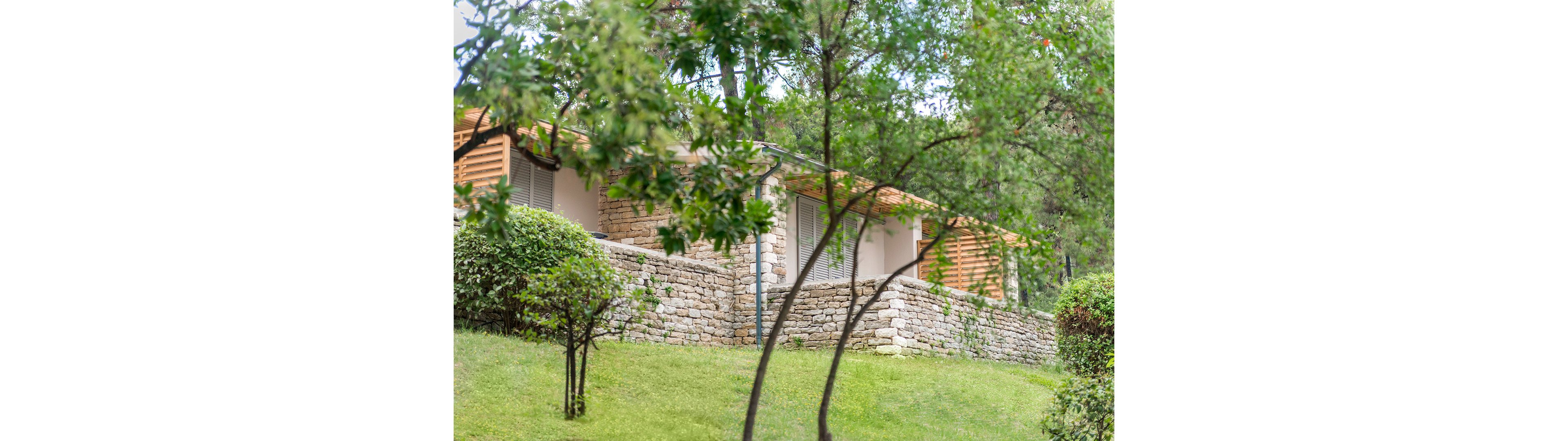 A low stone terrace house is partially visible behind lush green trees and shrubs. The building features light-colored stone walls, wooden slatted elements, and shuttered windows. It is set on a gently sloping grassy lawn, giving the scene a peaceful, natural atmosphere. Soft, diffused daylight enhances the greenery and the tranquil residential setting.